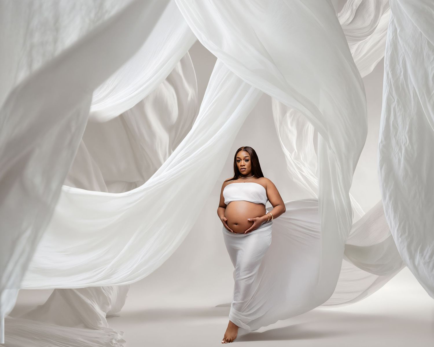 Pregnant woman in white crop top and skirt poses gracefully surrounded by flowing white fabric drapes in a bright studio.