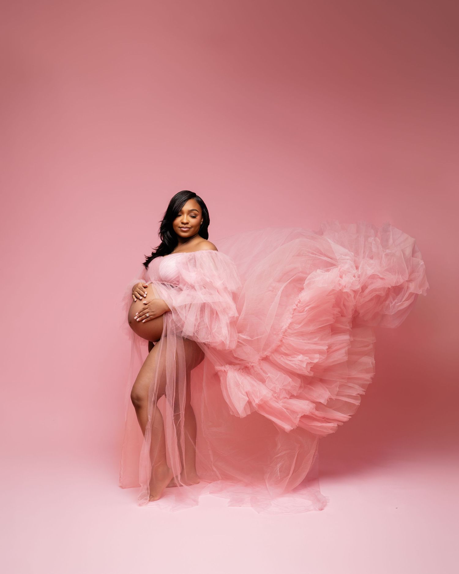 Pregnant woman in flowing pink tulle maternity gown poses gracefully against a soft pink studio background.