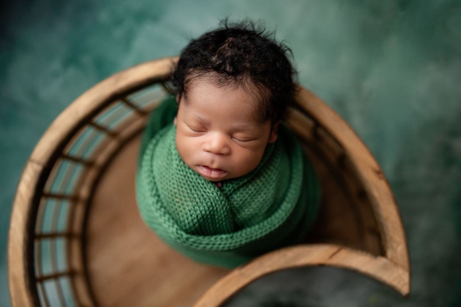 Newborn baby swaddled in green wrap sleeping peacefully inside a wooden basket on a teal background.