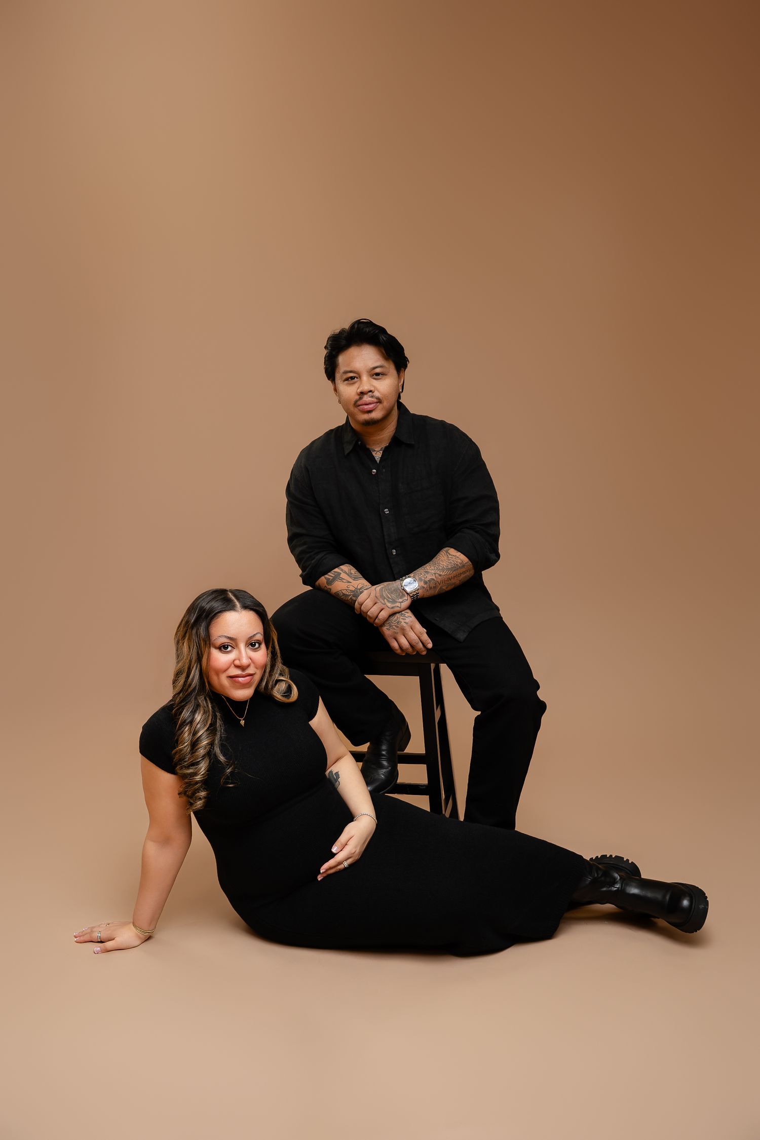 A pregnant woman in black sits on the floor beside a man on a stool, both dressed in black against a tan backdrop.