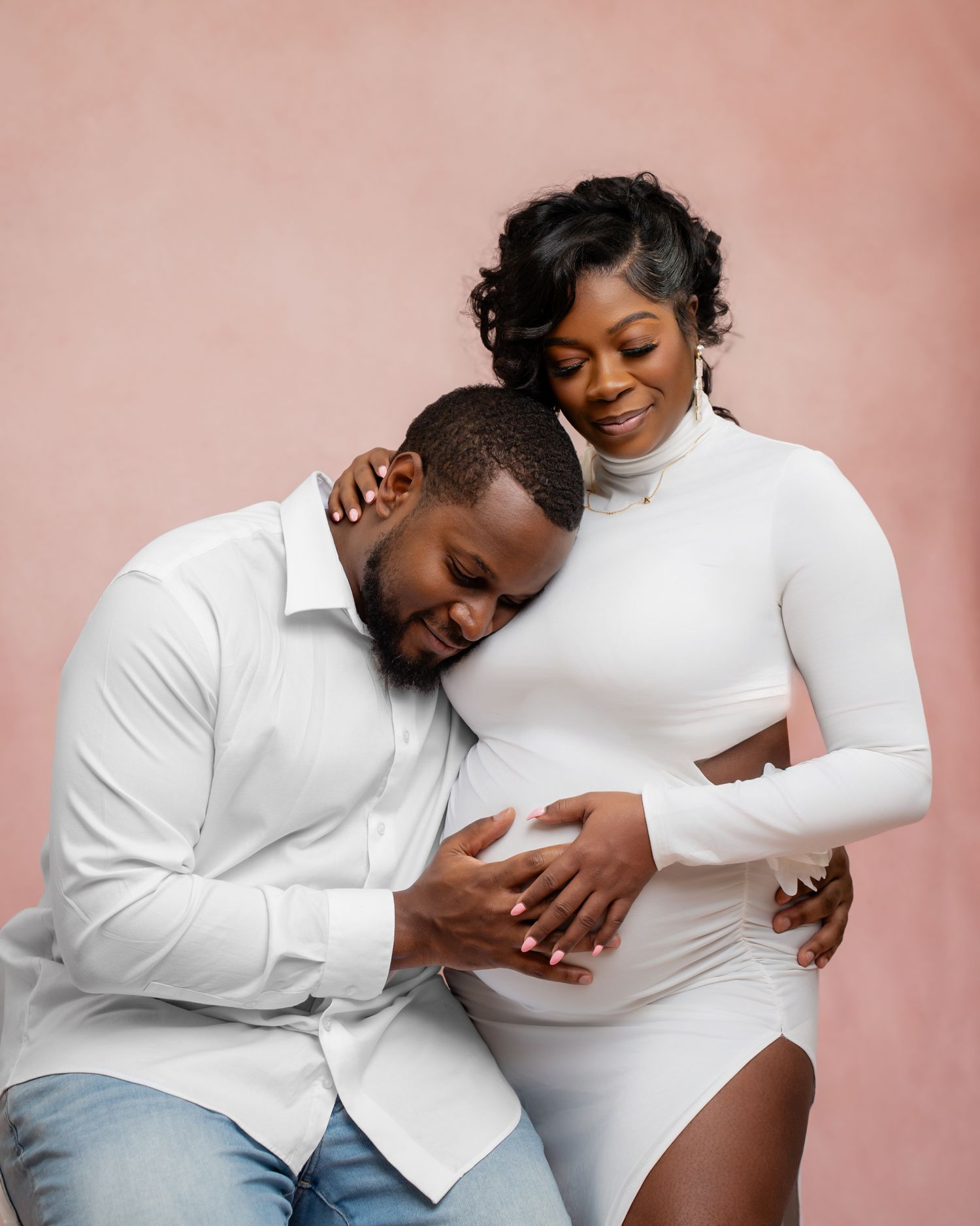 A couple in white outfits poses for a maternity photo, with the man tenderly resting his head on the pregnant woman's belly.