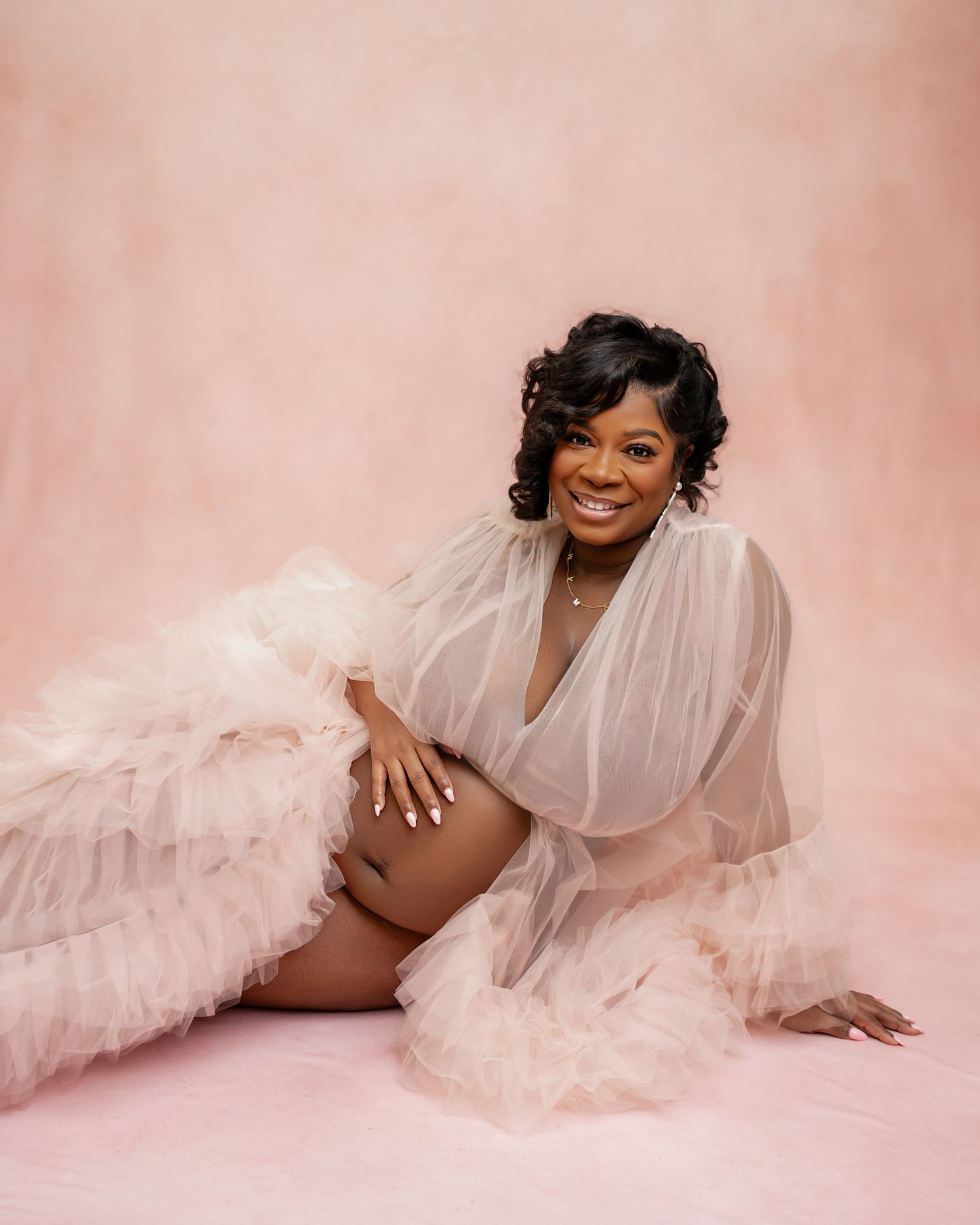 Smiling pregnant woman in a flowing pink tulle robe poses gracefully on a pink backdrop during a maternity photoshoot.