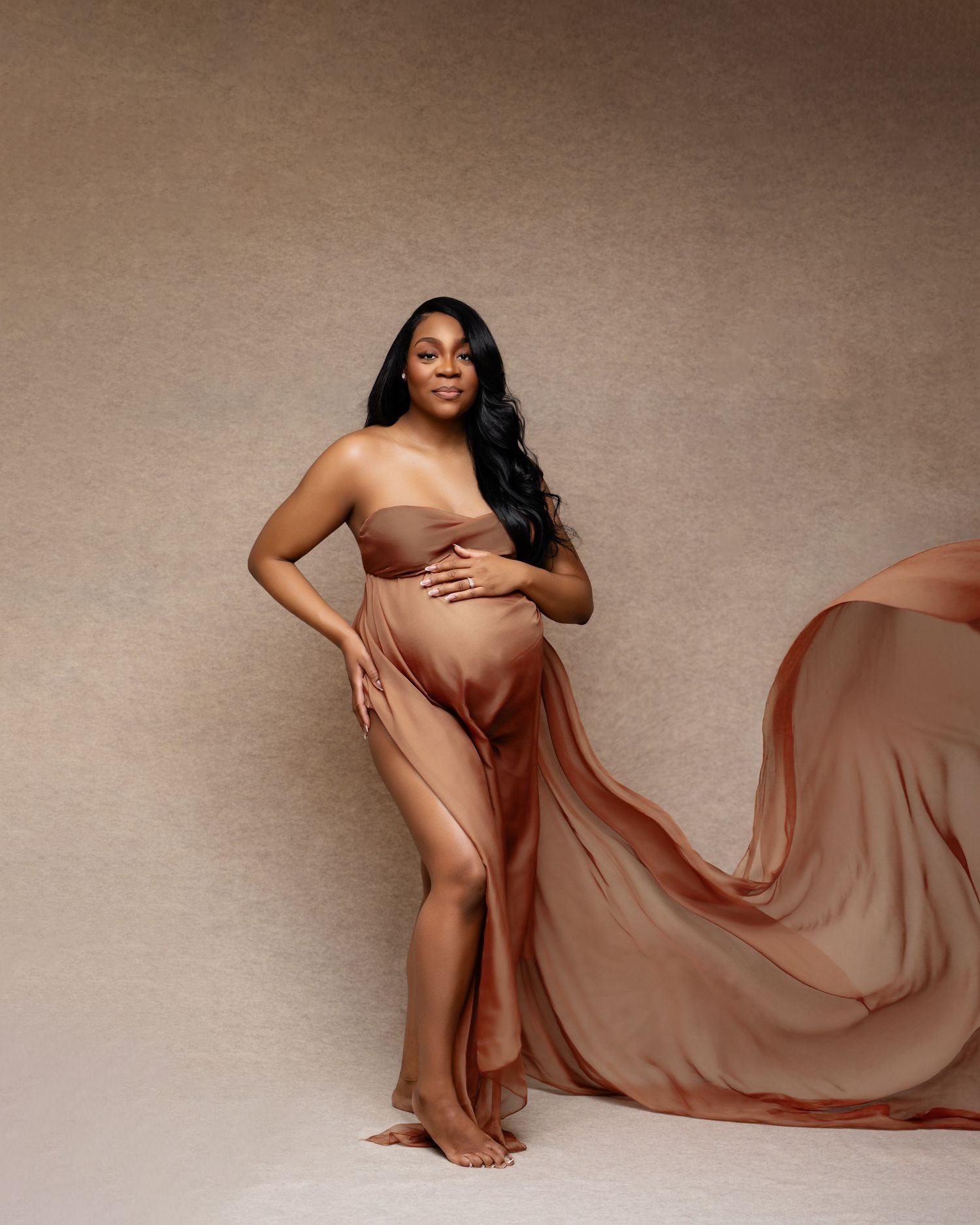 Pregnant woman in flowing terracotta chiffon maternity gown posing gracefully in a professional studio portrait.