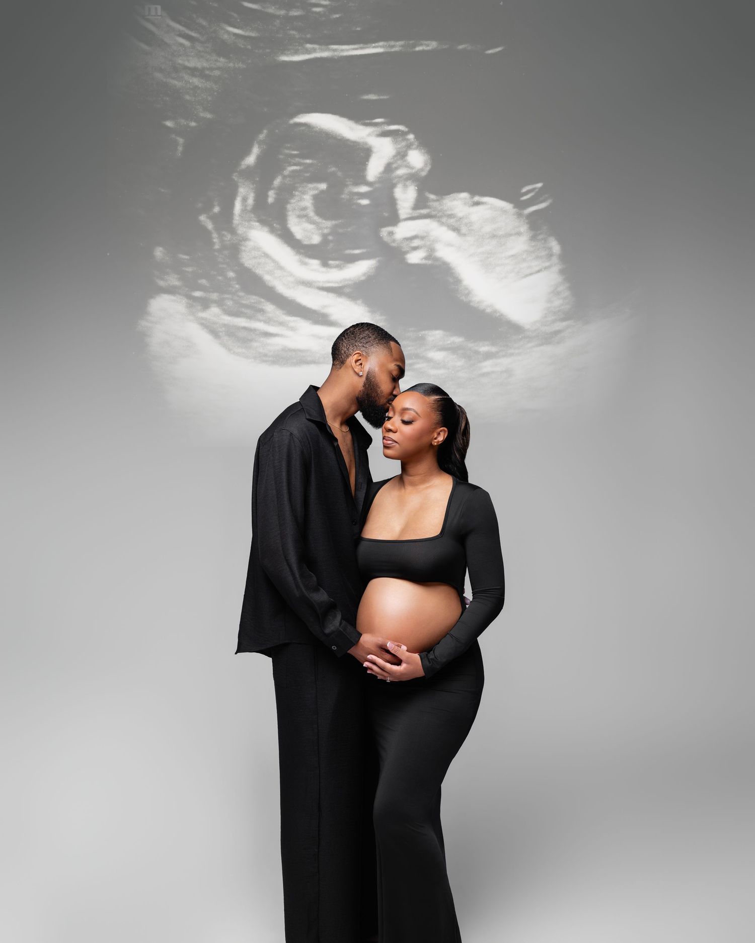 Couple in black maternity photoshoot with ultrasound projection backdrop, woman cradling baby bump.