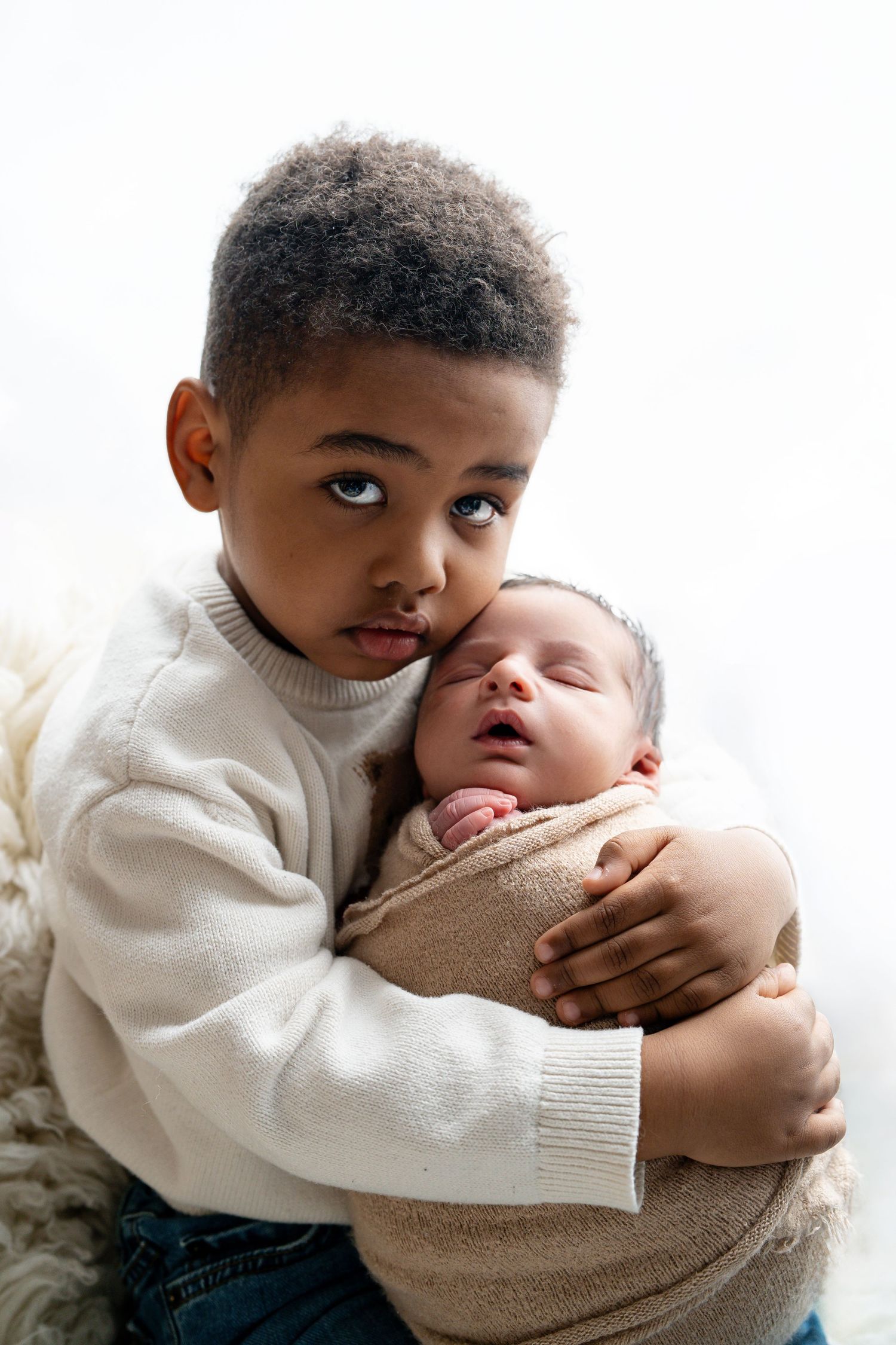 Young boy in white sweater gently holds sleeping newborn baby sibling, both dressed in neutral tones against white background.