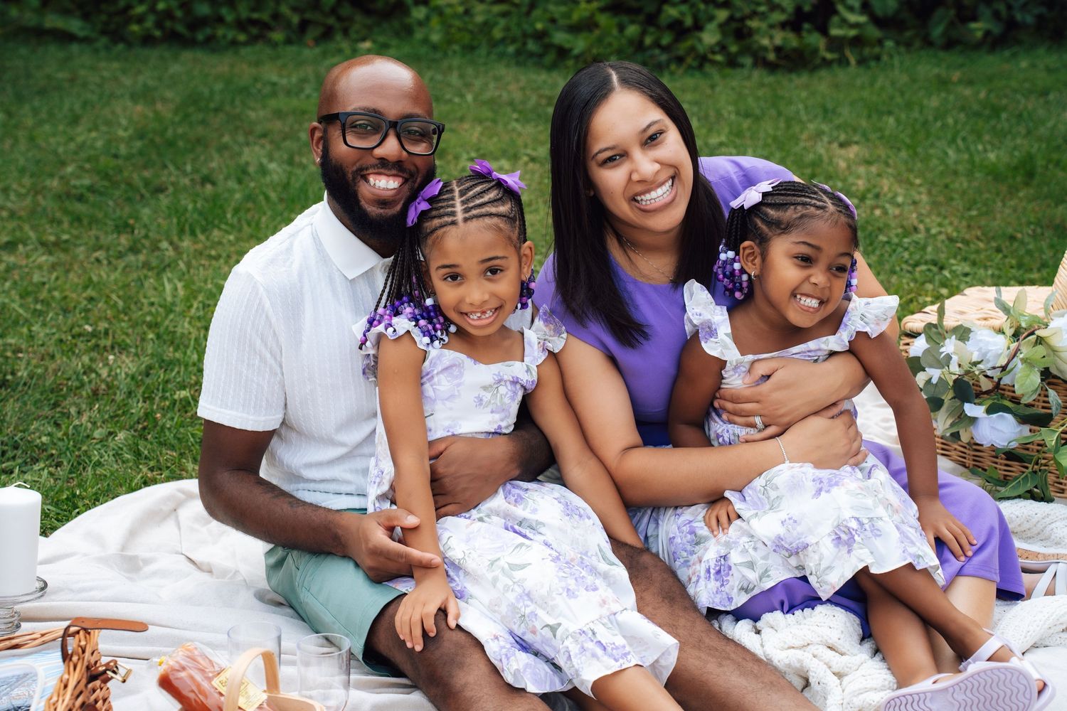 Metro Detroit family photographer capturing Family enjoying outdoor picnic on white blanket surrounded by lush green grass and trees.