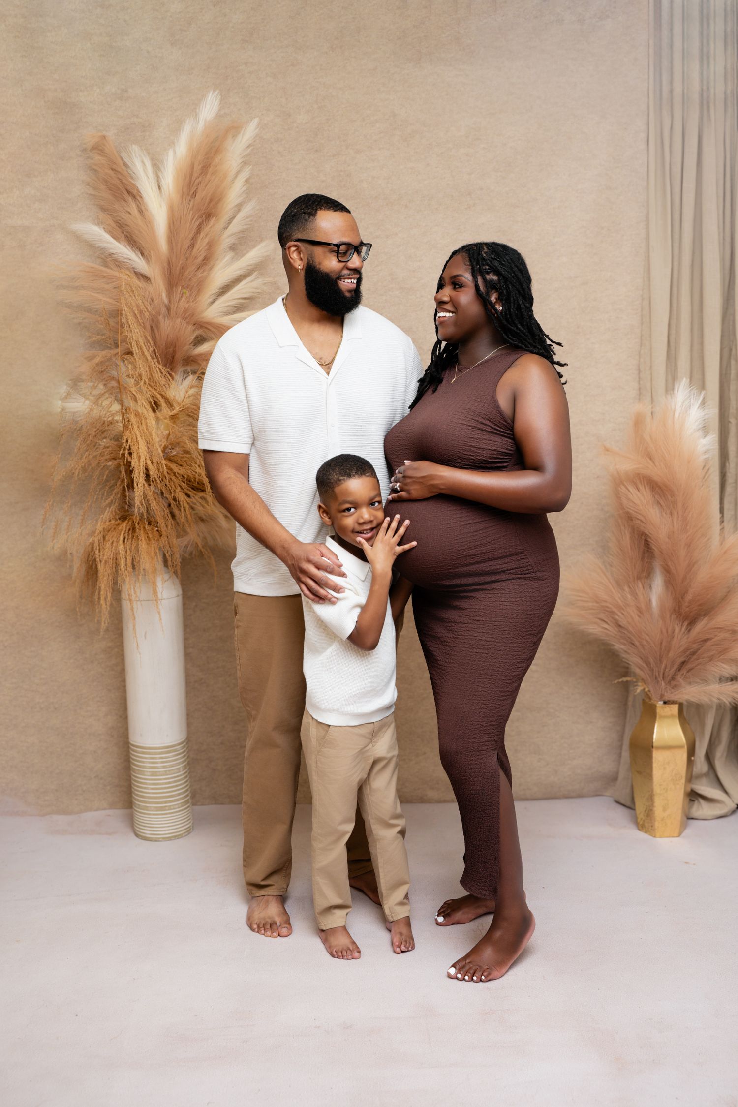 A happy pregnant Black family poses together in a studio with pampas grass decor, featuring a man, woman, and young son.