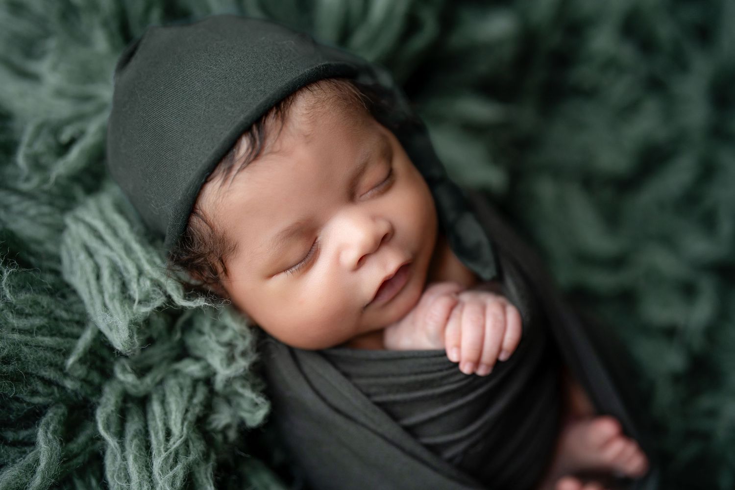 Newborn baby sleeping peacefully wrapped in dark green swaddle with matching knit hat on fluffy green backdrop.