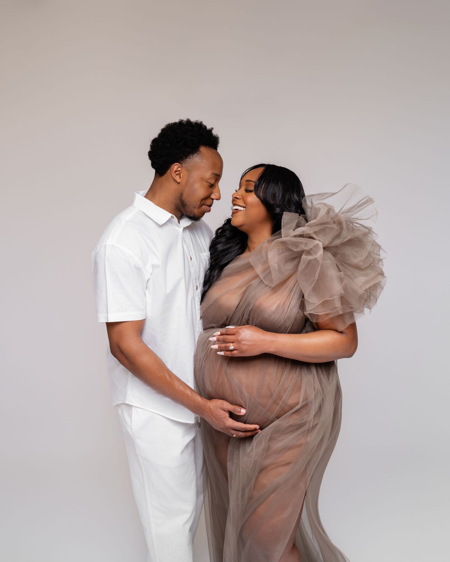 A pregnant couple poses tenderly in a studio, man in white outfit, woman in sheer taupe gown cradling her baby bump.
