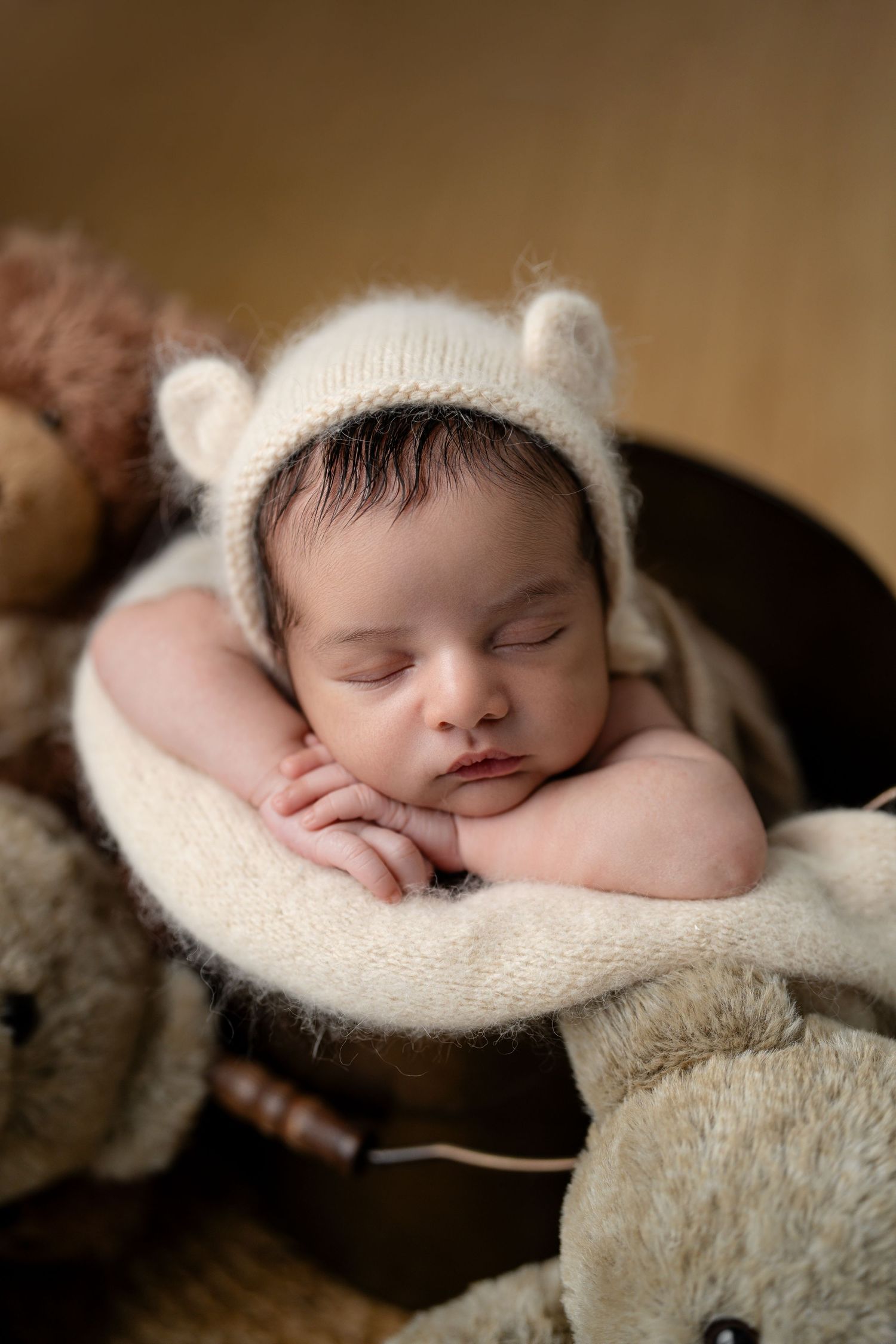 Sleeping newborn baby wearing a cream knit bear bonnet hat, resting in a wooden bucket surrounded by soft teddy bears.