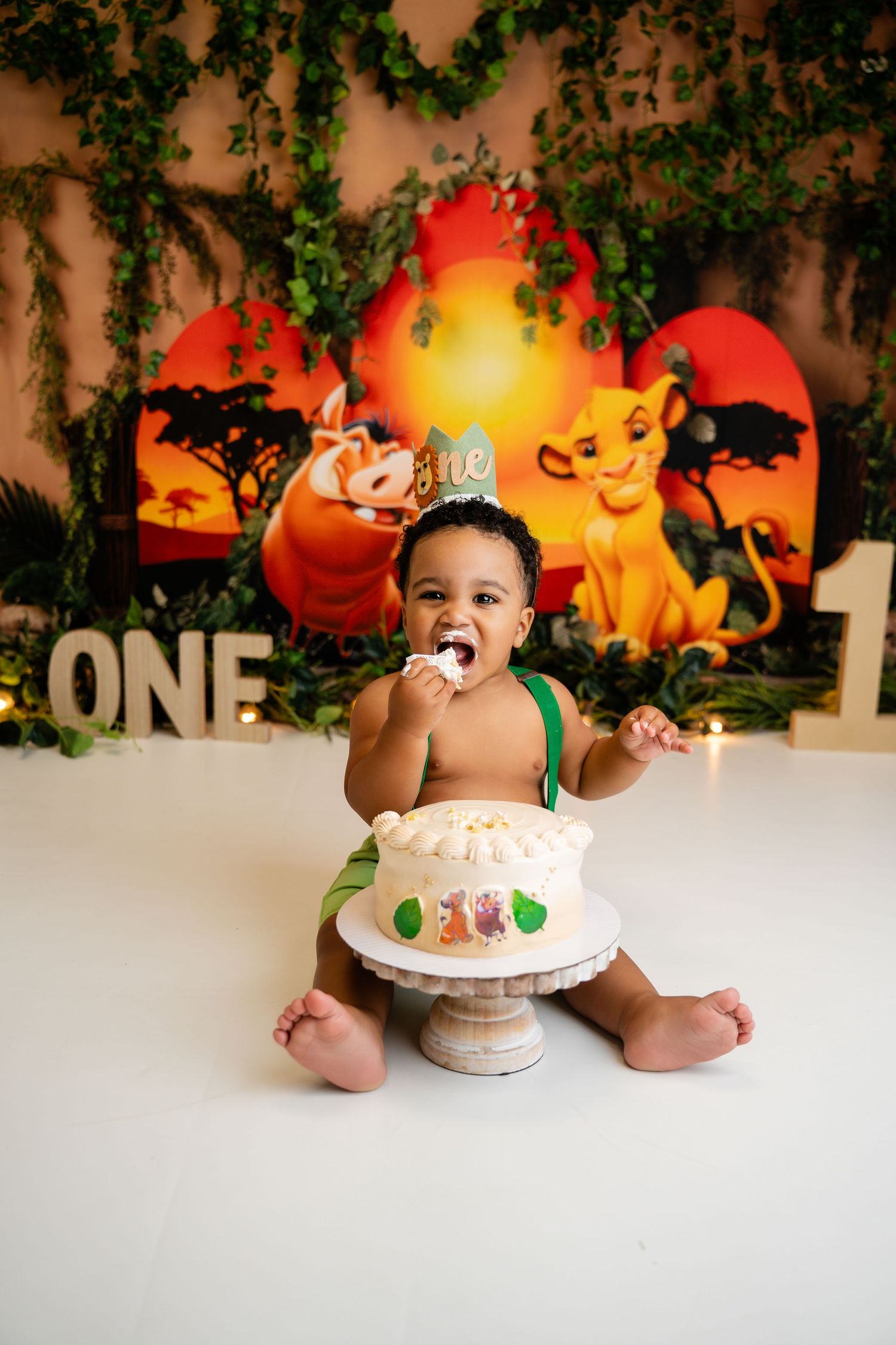 Baby boy celebrates first birthday with Lion King themed cake smash, wearing green outfit and gold crown.