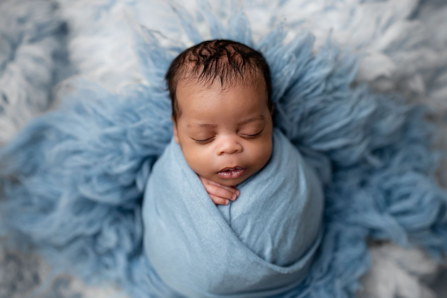 Newborn baby swaddled in blue wrap sleeping peacefully on a fluffy blue fur blanket in a professional photo.