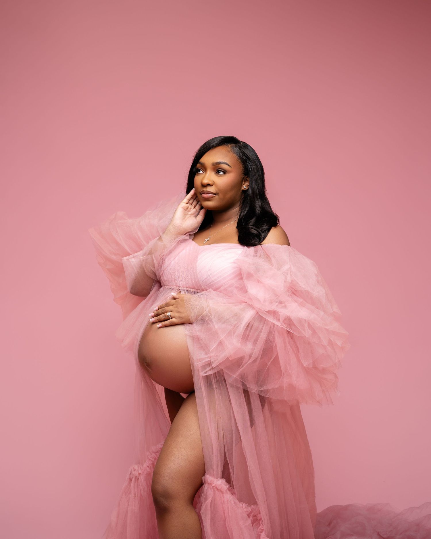 Pregnant woman in pink tulle maternity gown poses gracefully against a pink studio background.