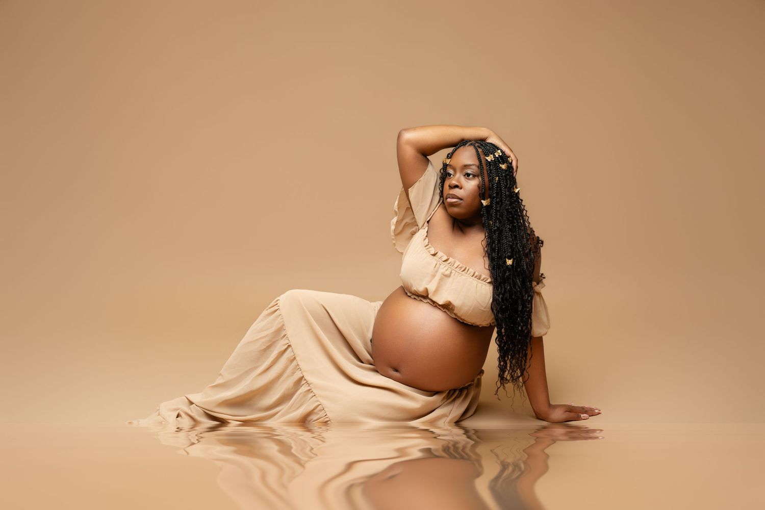 Pregnant woman in beige crop top and flowing skirt poses gracefully on reflective studio floor with braided hair.