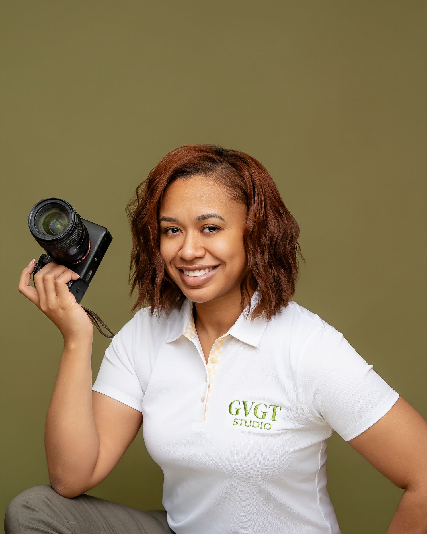 Professional photographer in white polo shirt with company logo holding DSLR camera against olive green background.