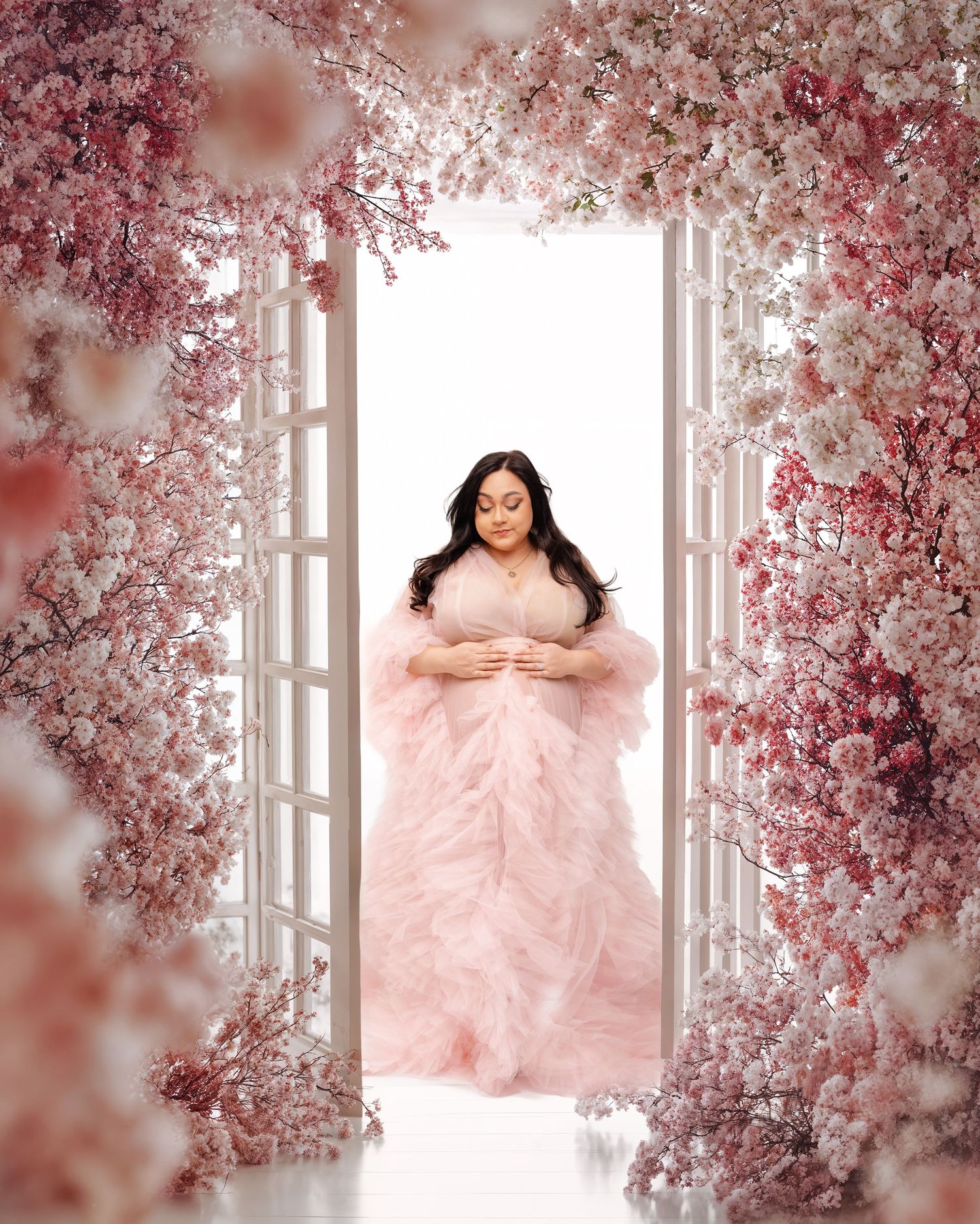 Pregnant woman in pink tulle gown stands in floral doorway surrounded by cherry blossoms in elegant maternity photo.
