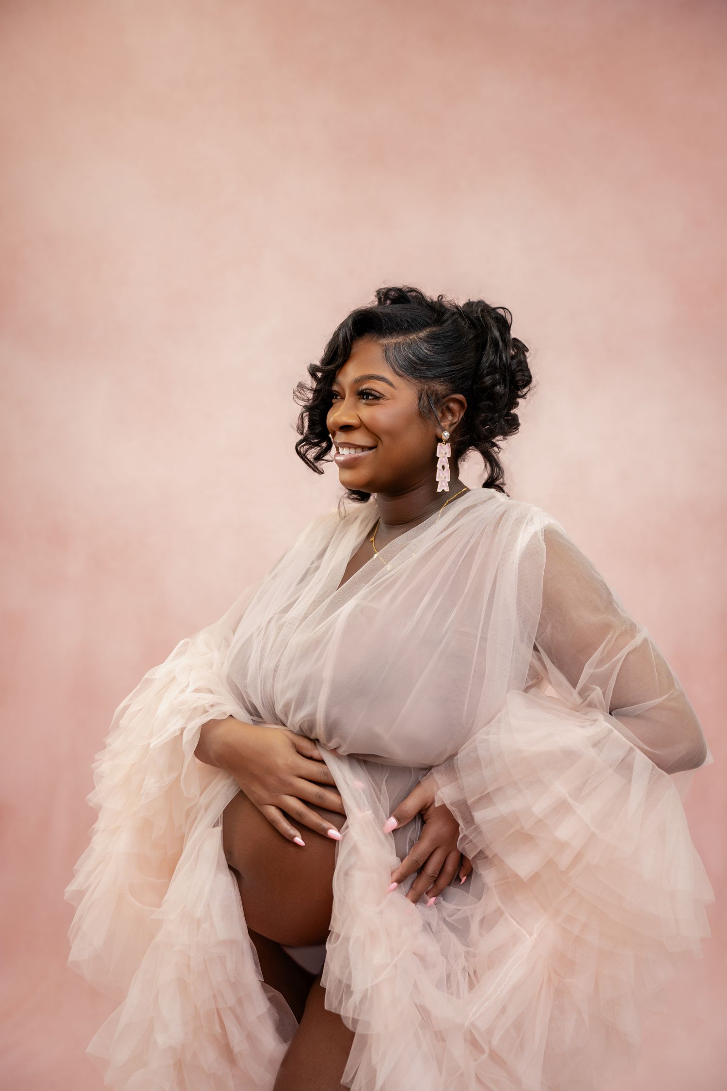 Smiling pregnant woman in sheer blush robe poses against pink backdrop in elegant maternity portrait.