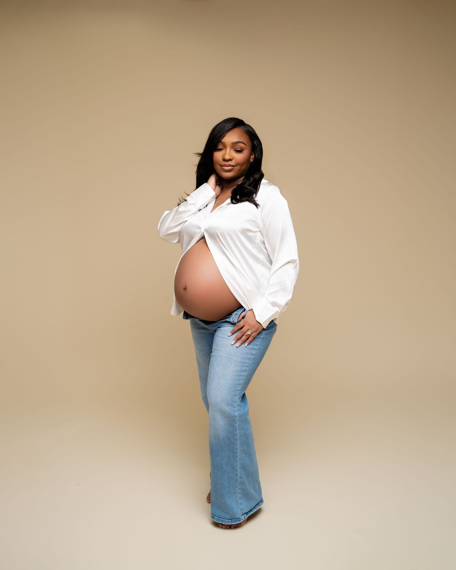 Pregnant woman in white shirt and flare jeans posing for maternity photo on beige background.