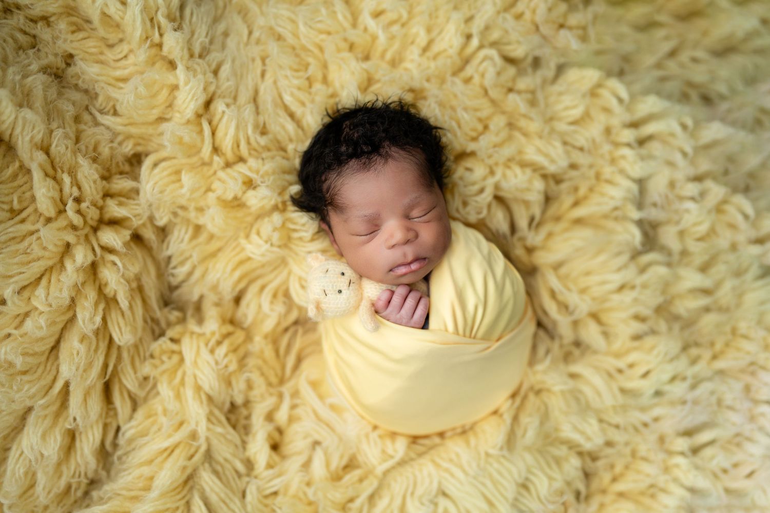Newborn baby swaddled in yellow wrap sleeping peacefully on a soft fluffy yellow blanket with a tiny teddy bear.