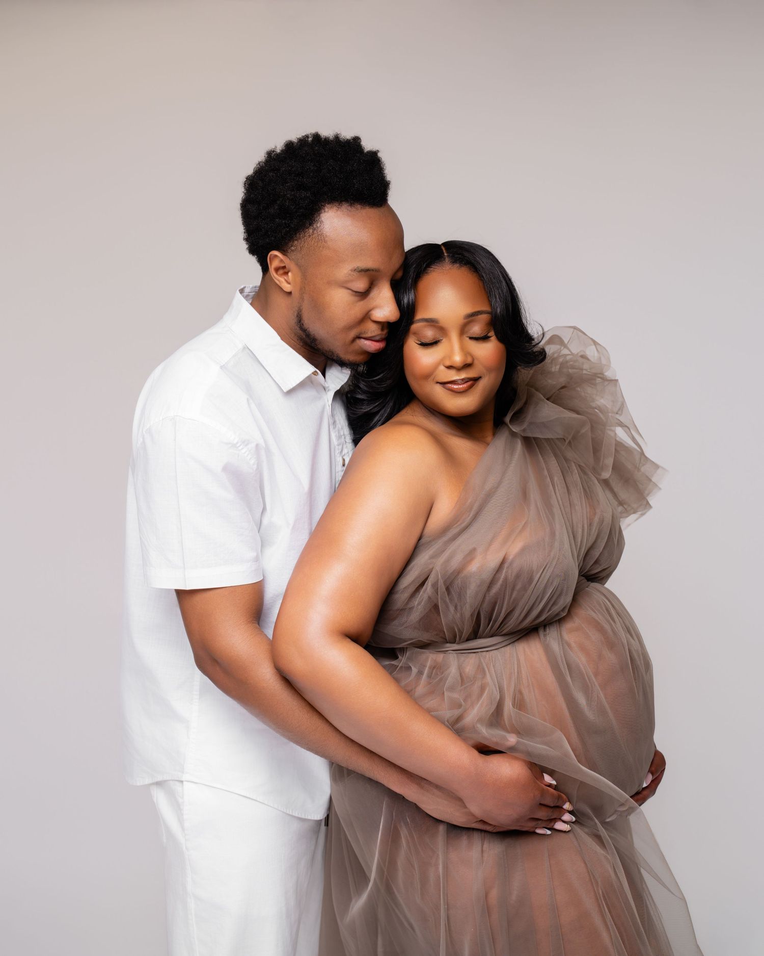 A couple poses for a maternity photo, the pregnant woman in a sheer taupe gown cradled by her partner in white.