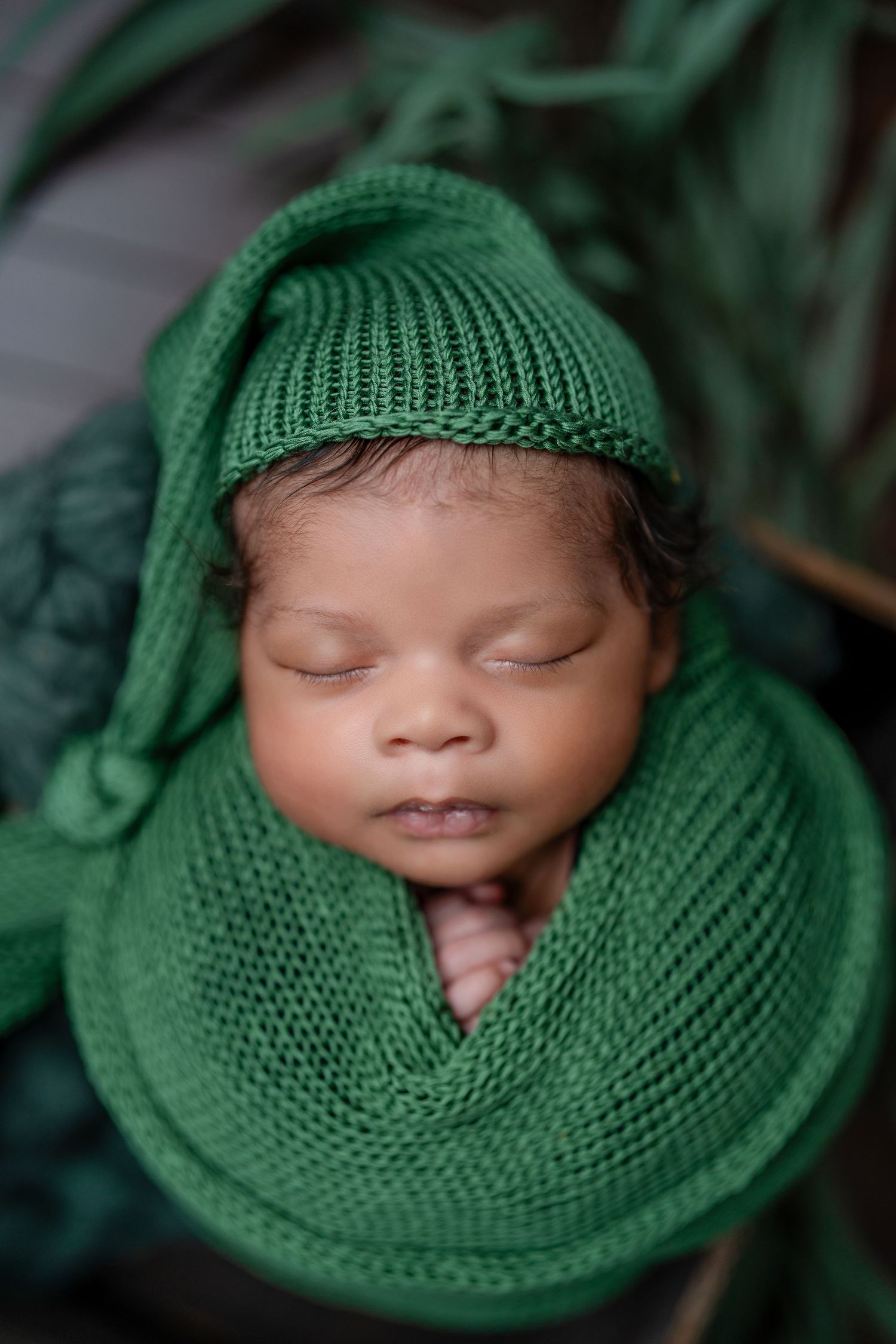 Sleeping newborn baby wrapped in a green knit blanket and matching hat, peacefully resting in a cozy basket.