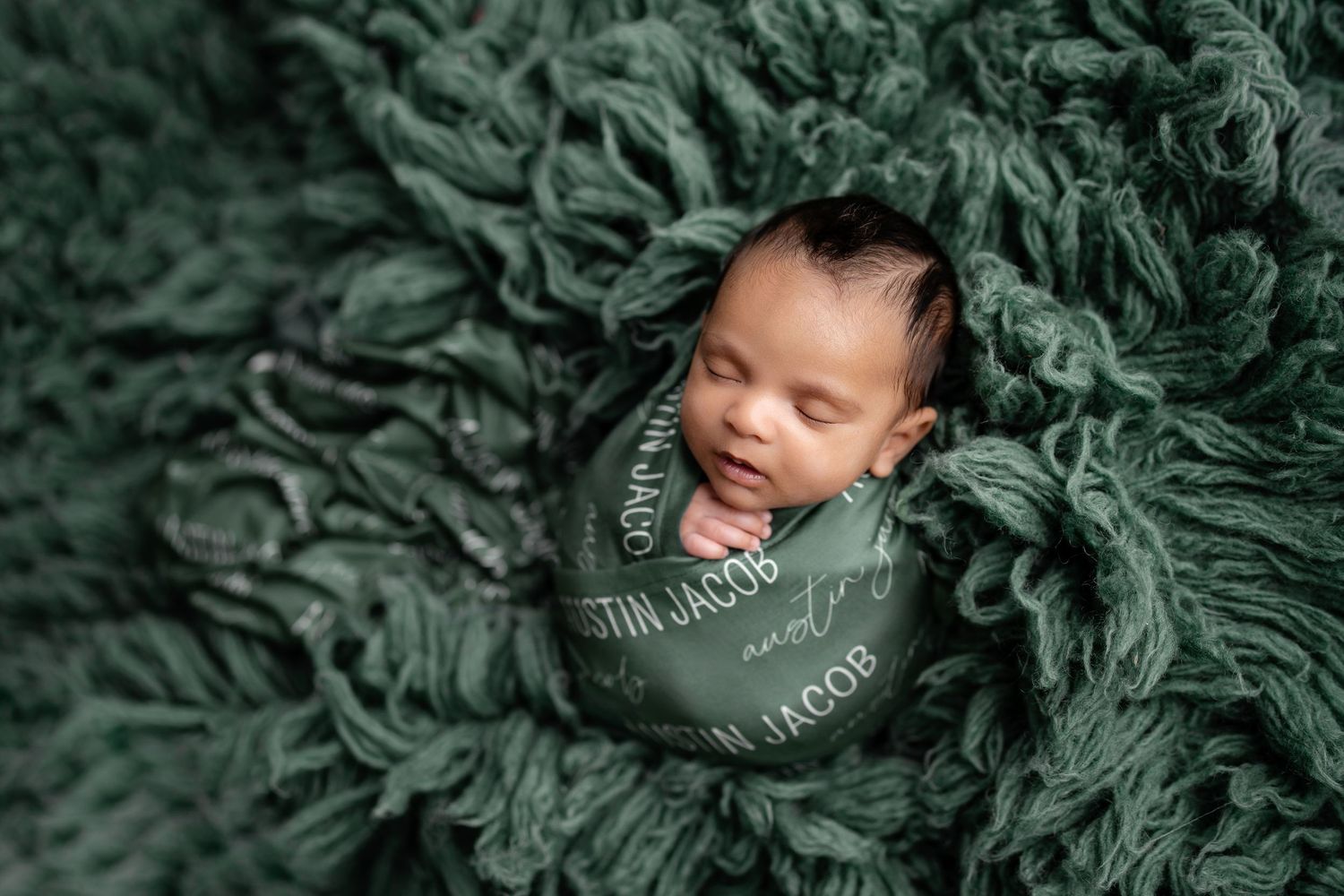 Newborn baby swaddled in a green personalized wrap blanket, resting peacefully on a fluffy dark green textured rug.