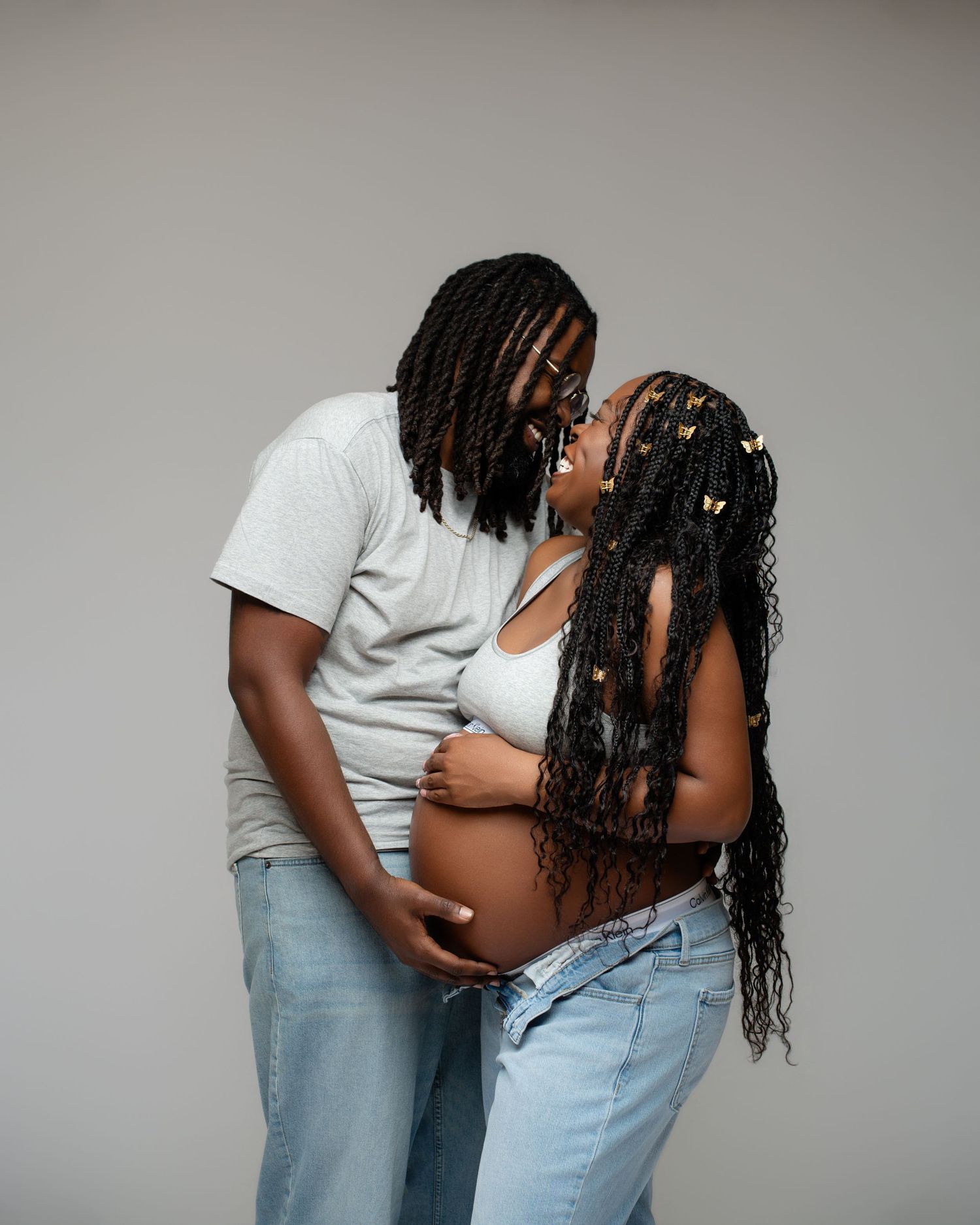 A couple shares a tender moment during a maternity photoshoot, both wearing casual denim, with butterfly hair accessories.