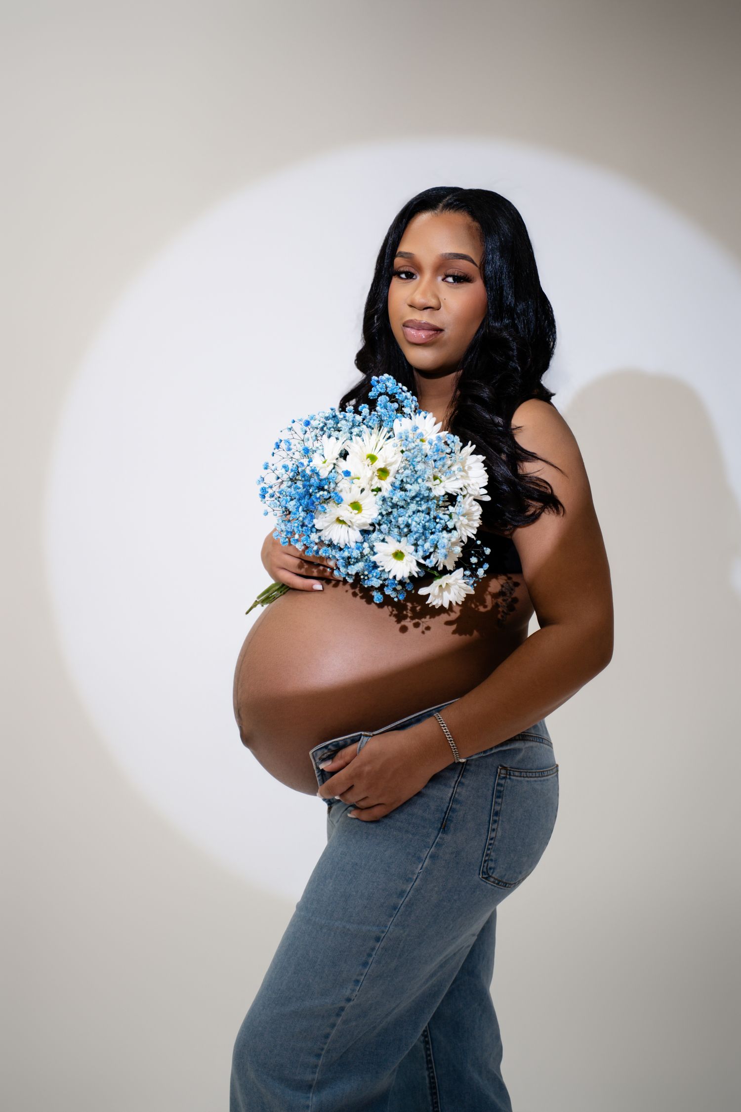 Pregnant woman holding blue and white flowers bouquet, wearing jeans, posing in studio with spotlight background.