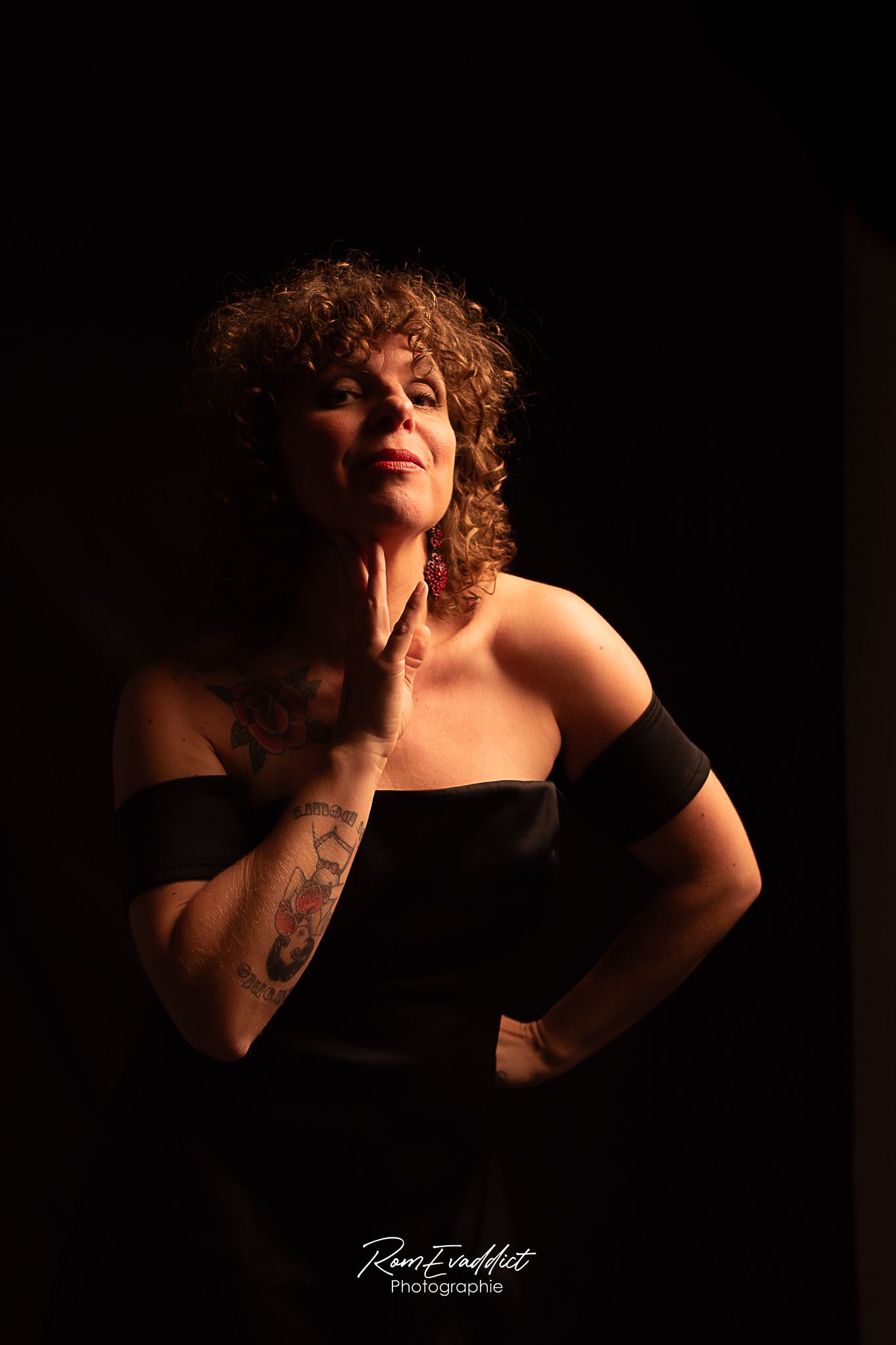 femme cheveux bouclés robe noir tatouage sur l'avant bras se tenant debout la main posée sur la joue-portrait boudoir intimiste