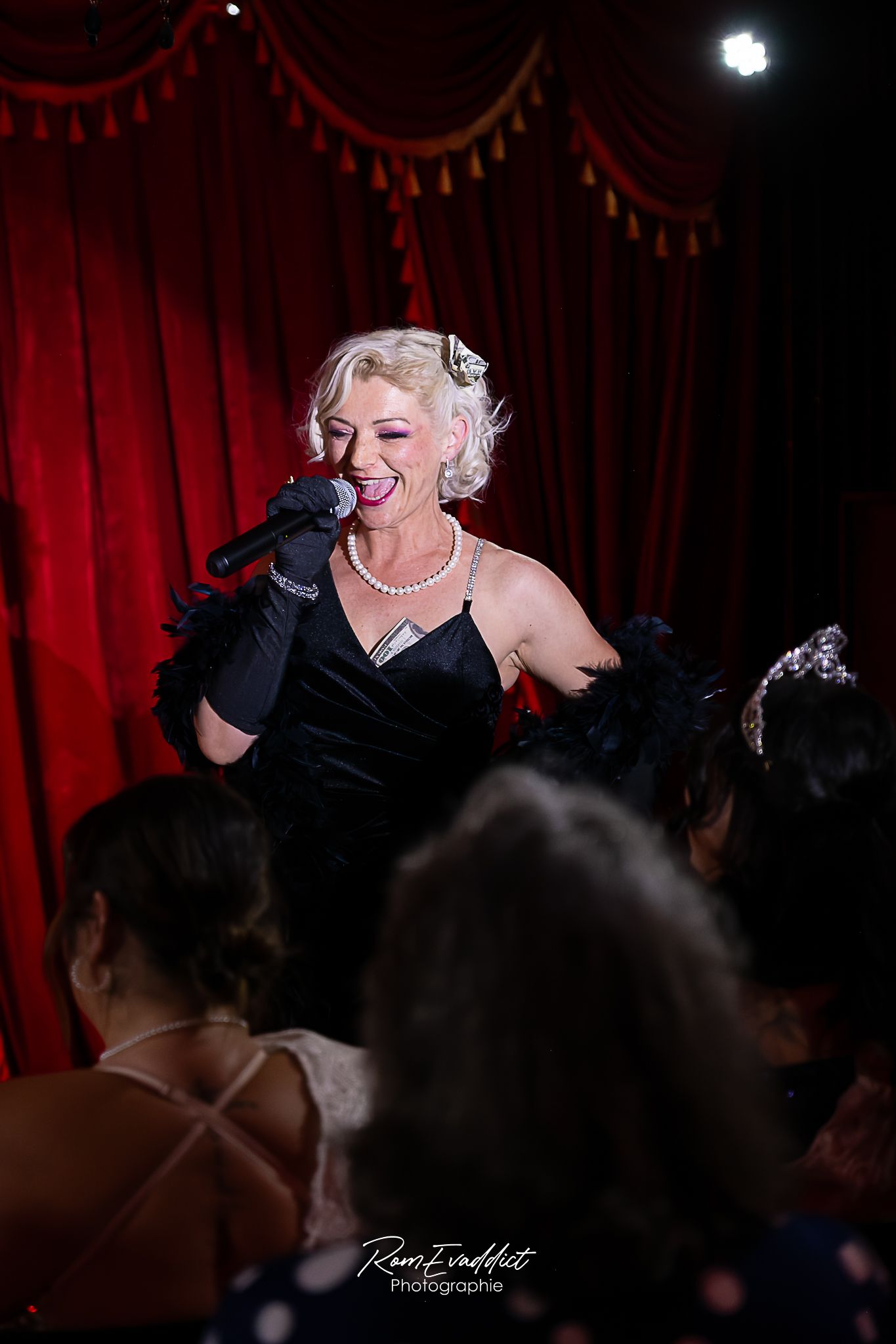 photo chanteuse de cabaret-blonde robe longue à strass noirs- chantant dans le public durant spectacle de cabaret- fond rideau rouge