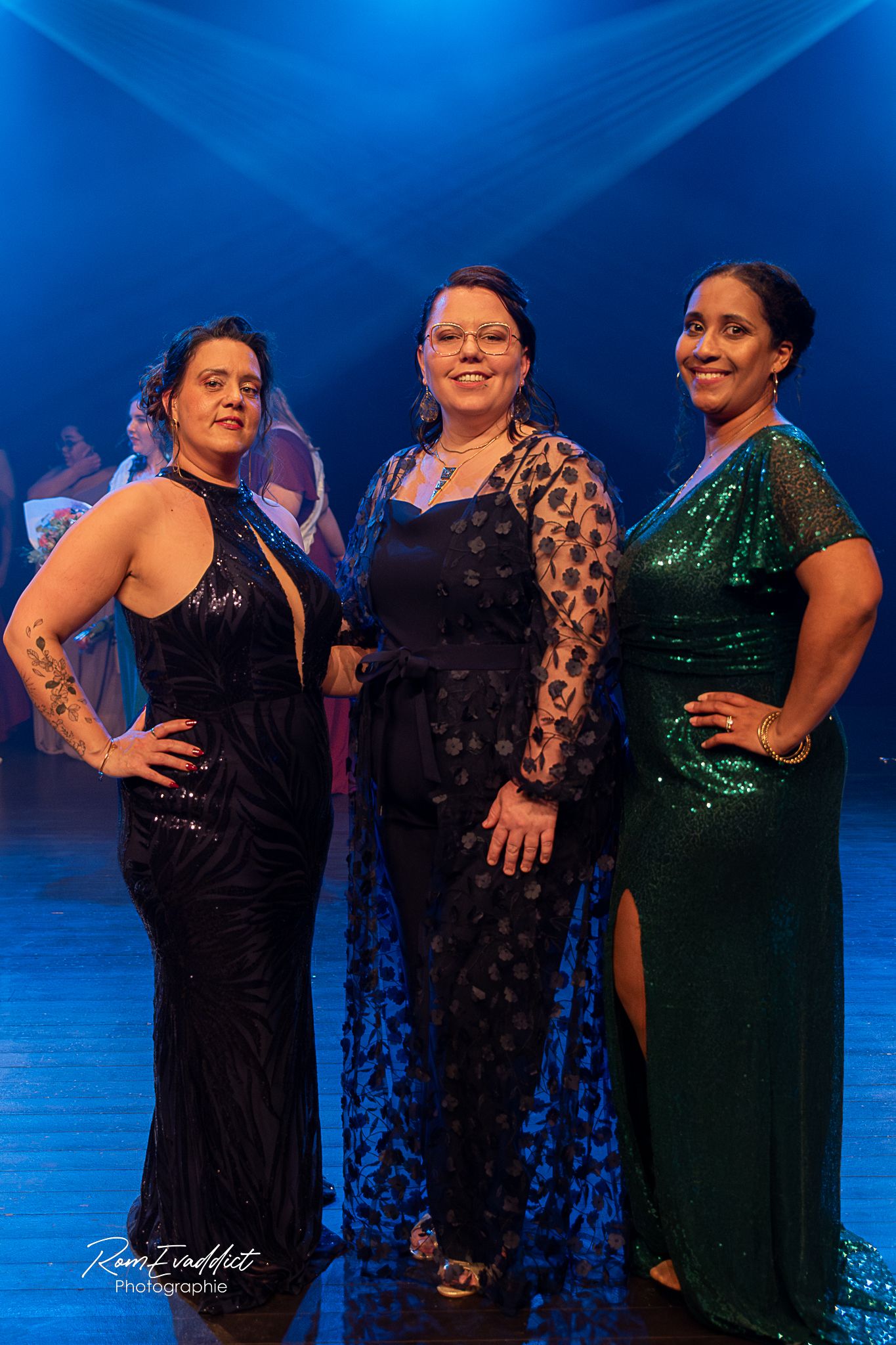3 femmes se tenant debout sur scène en robe de soirée, une noire, une bleu une verte, éclairage bleu- événementiel