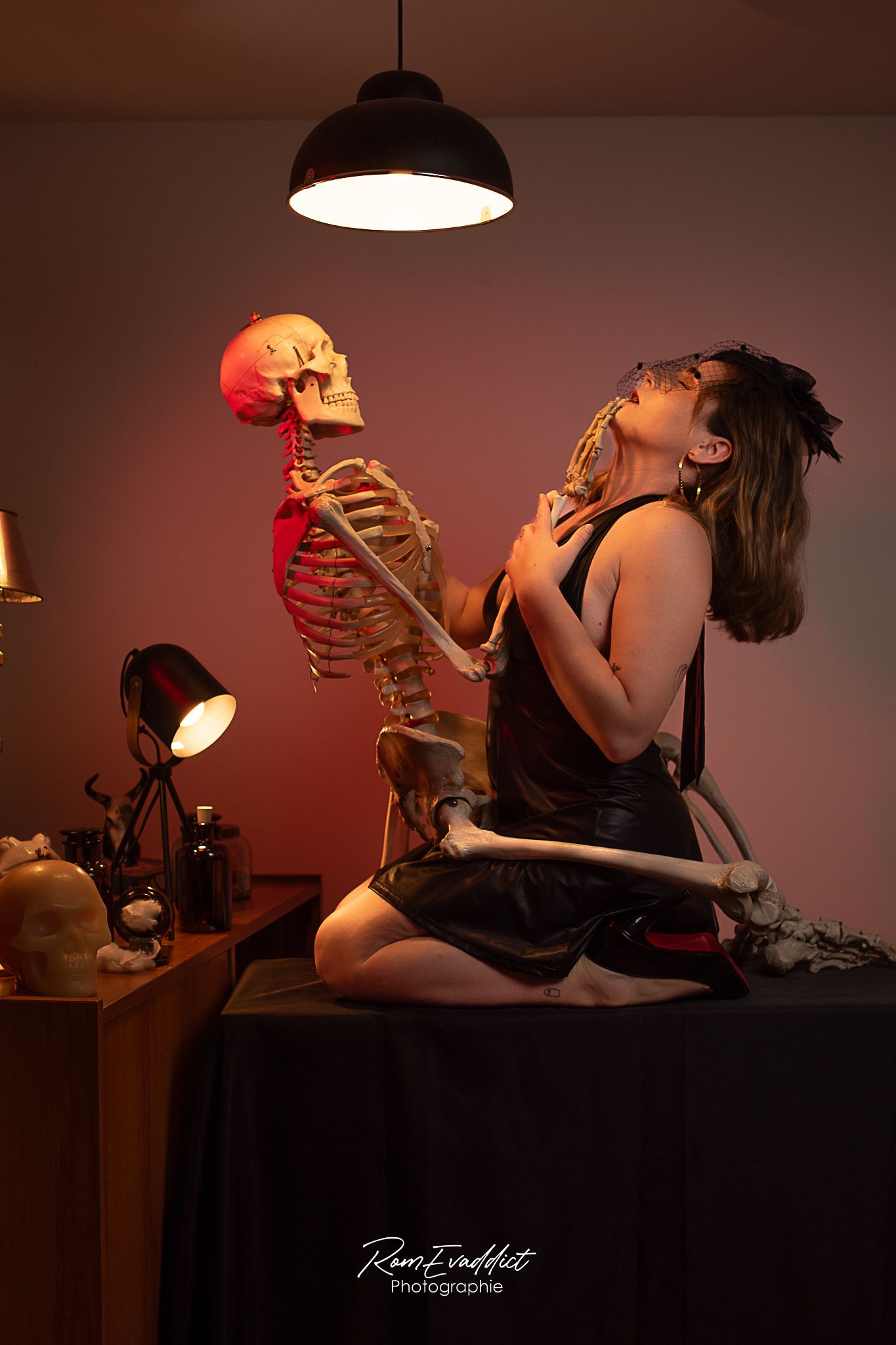 femme robe noir assise sur une table noir enlacée avec un squelette-ambiance cabinet de curiosité lumière rouge-portrait boudoir intimiste