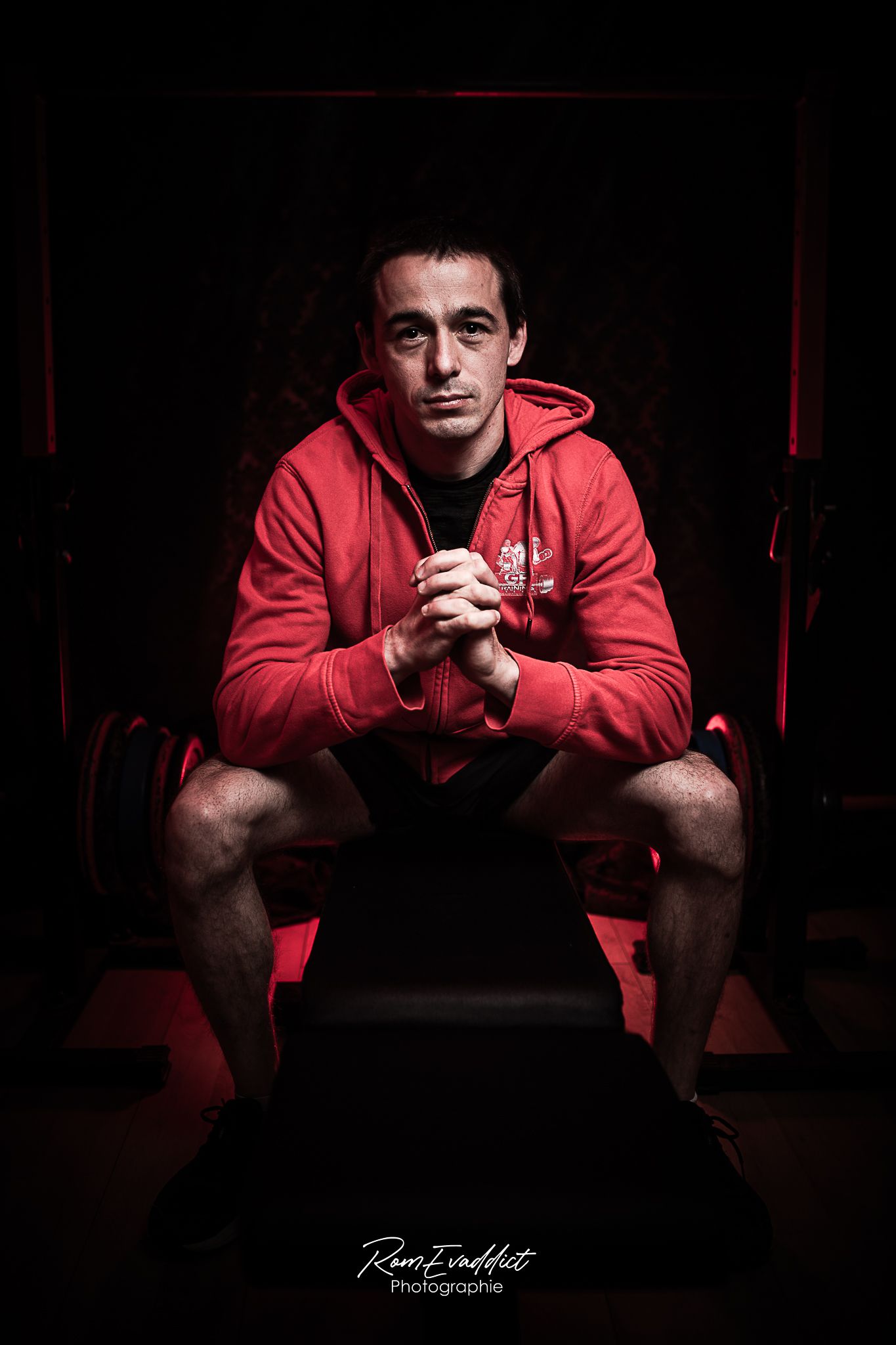 homme coach sportif en short noir et sweat rouge assis sur banc d'abdo faisant face caméra pour photo portrait corporate d'entreprise