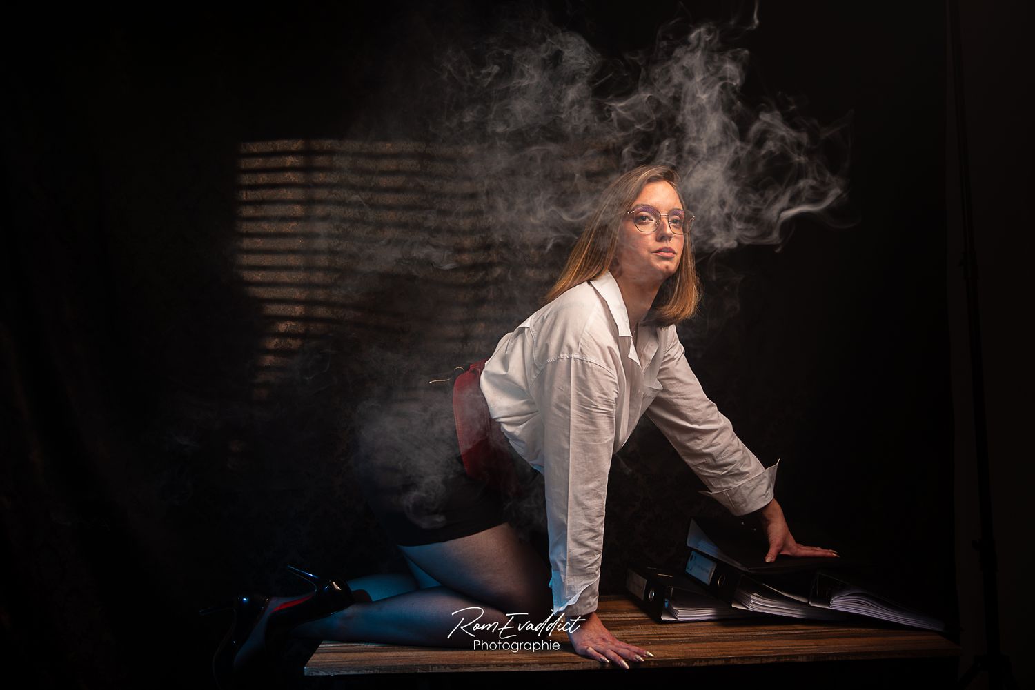 femme en jupe crayon noir chemisier blanc à quatre pattes sur un bureau peu éclairé-fumée-ombres-portrait boudoir confiance en soi
