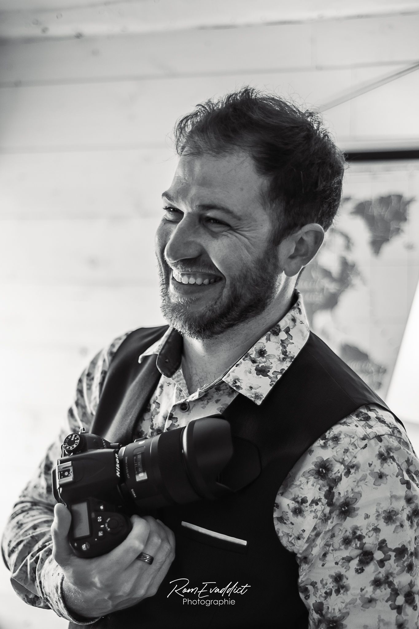 photo noir et blanc homme souriant en gilet bleu et chemise blanche à petite fleurs tenant un appareil photo à la main-portrait boudoir