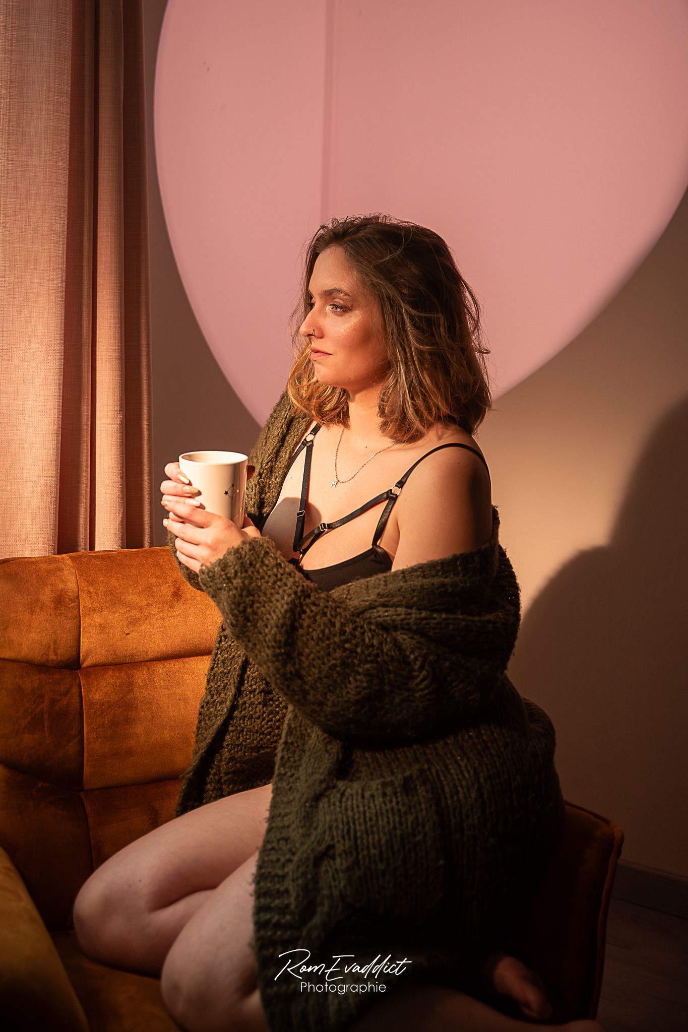 femme en body noir et pull vert olive à genou dans un fauteuil une tasse fumante à la main-éclairage cinéma-portrait boudoir intimiste