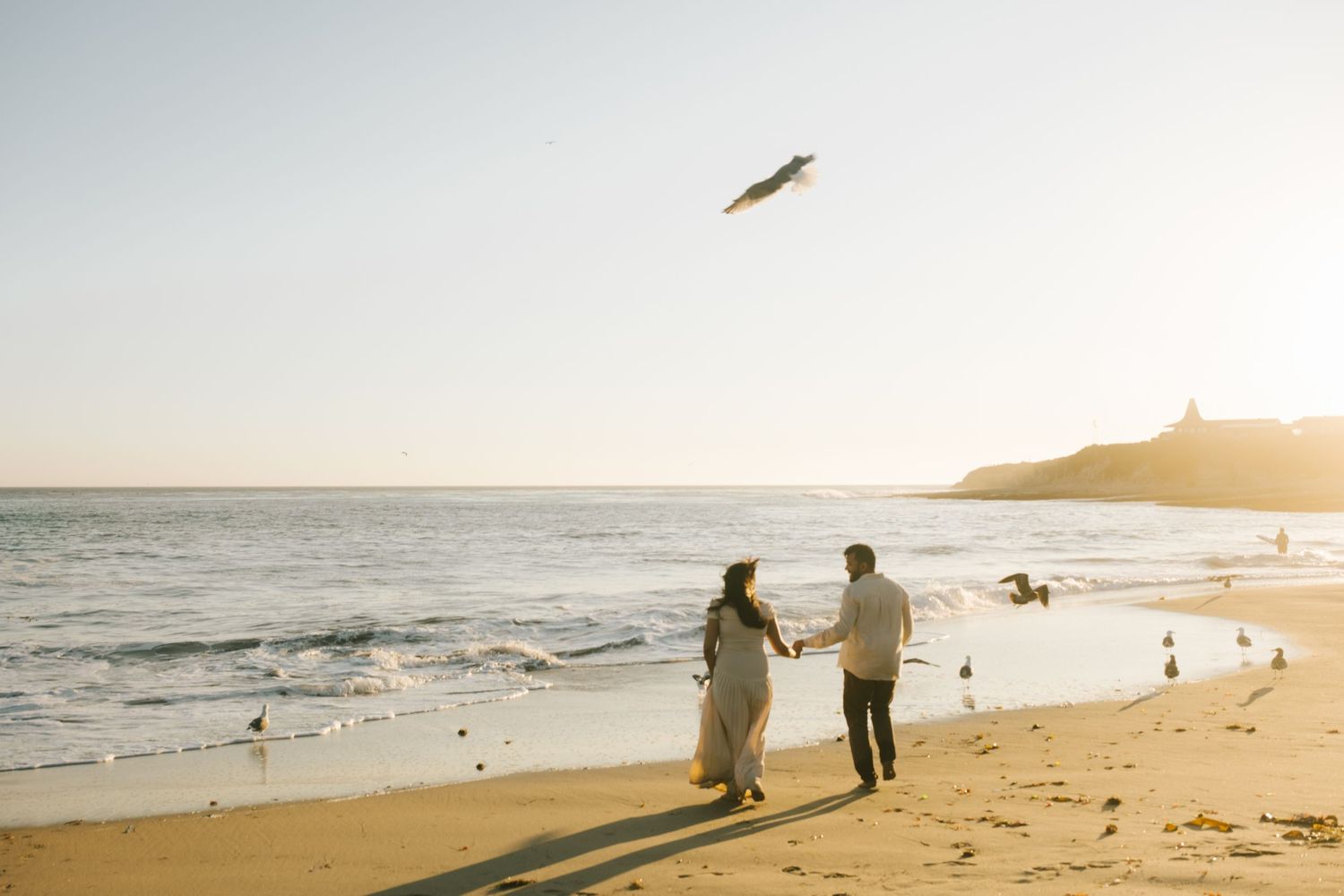 Silhouettes of people walking along beach at sunset with lighthouse in the distance.