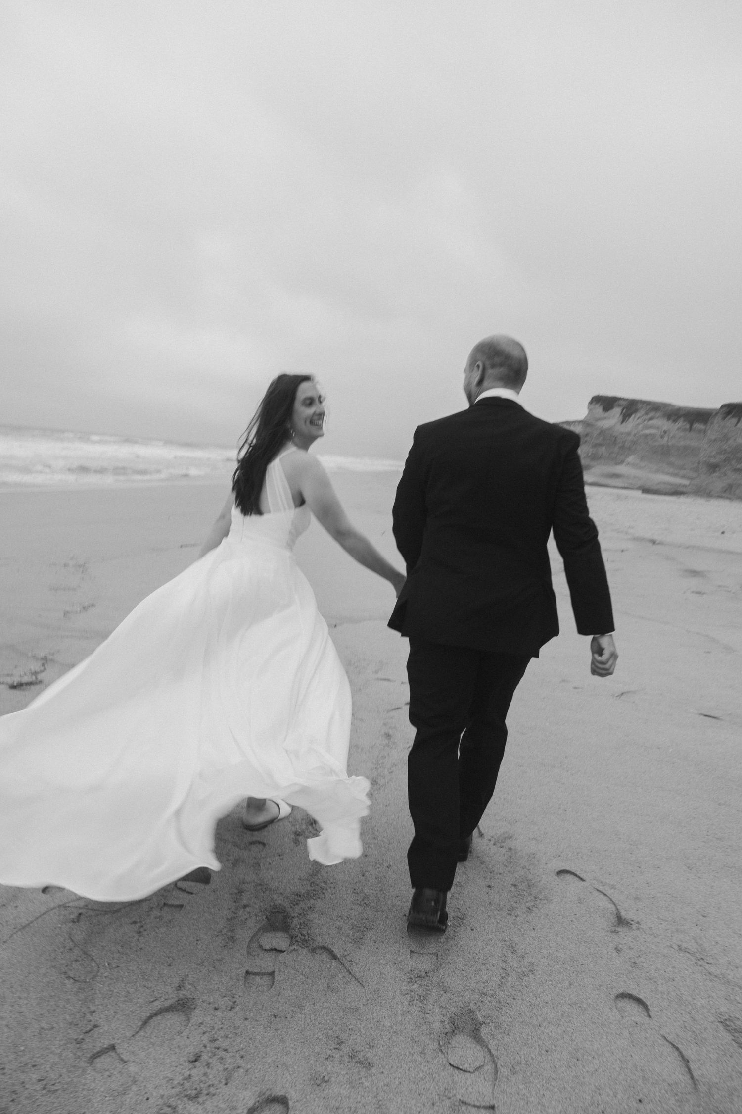 A bride and groom walking hand in hand on a beach, her dress flowing in the wind.
