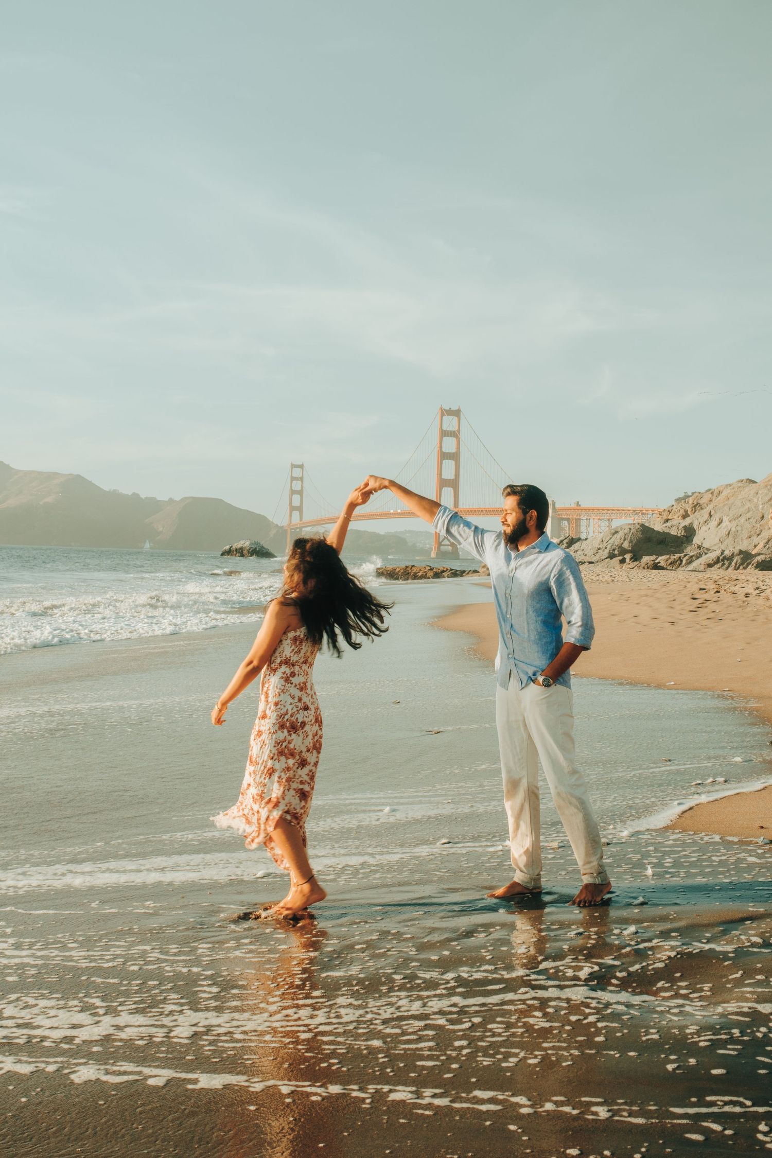 Playful couple dancing on beach with Golden Gate Bridge in background.