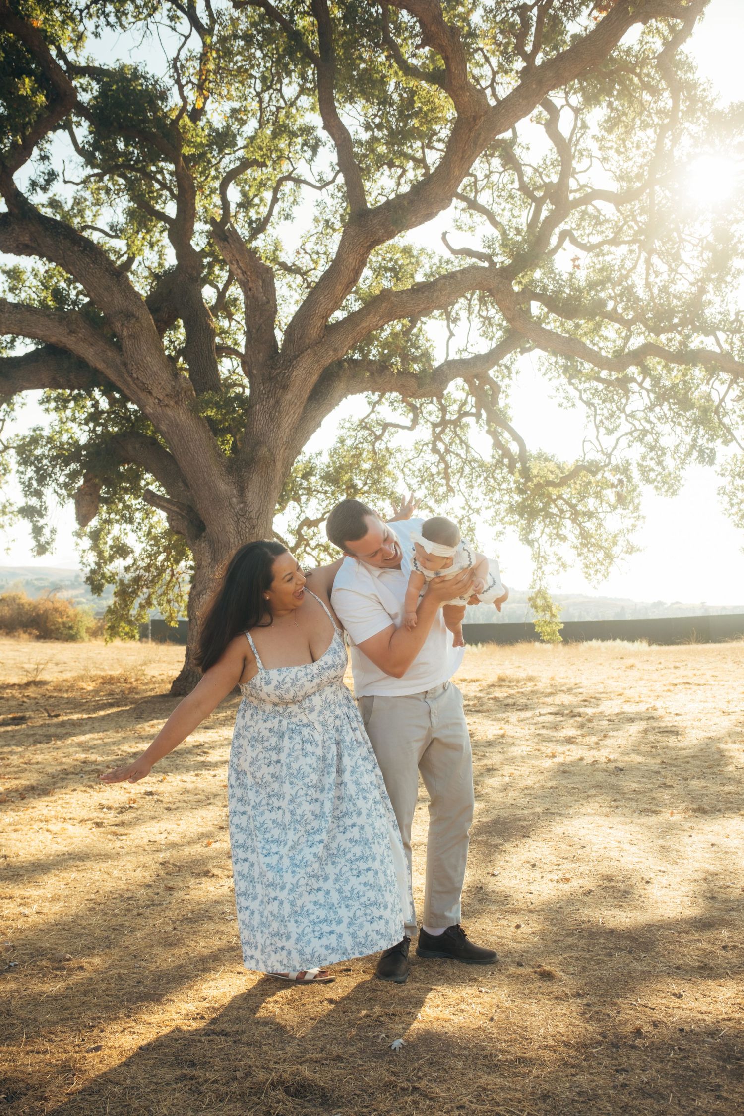 Family shares playful moment under large oak tree during golden hour.