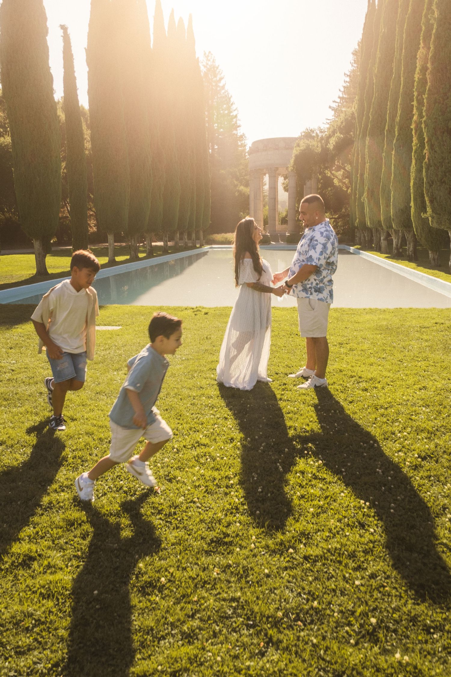 Family members playing and running together on a sunlit grassy lawn with long shadows.