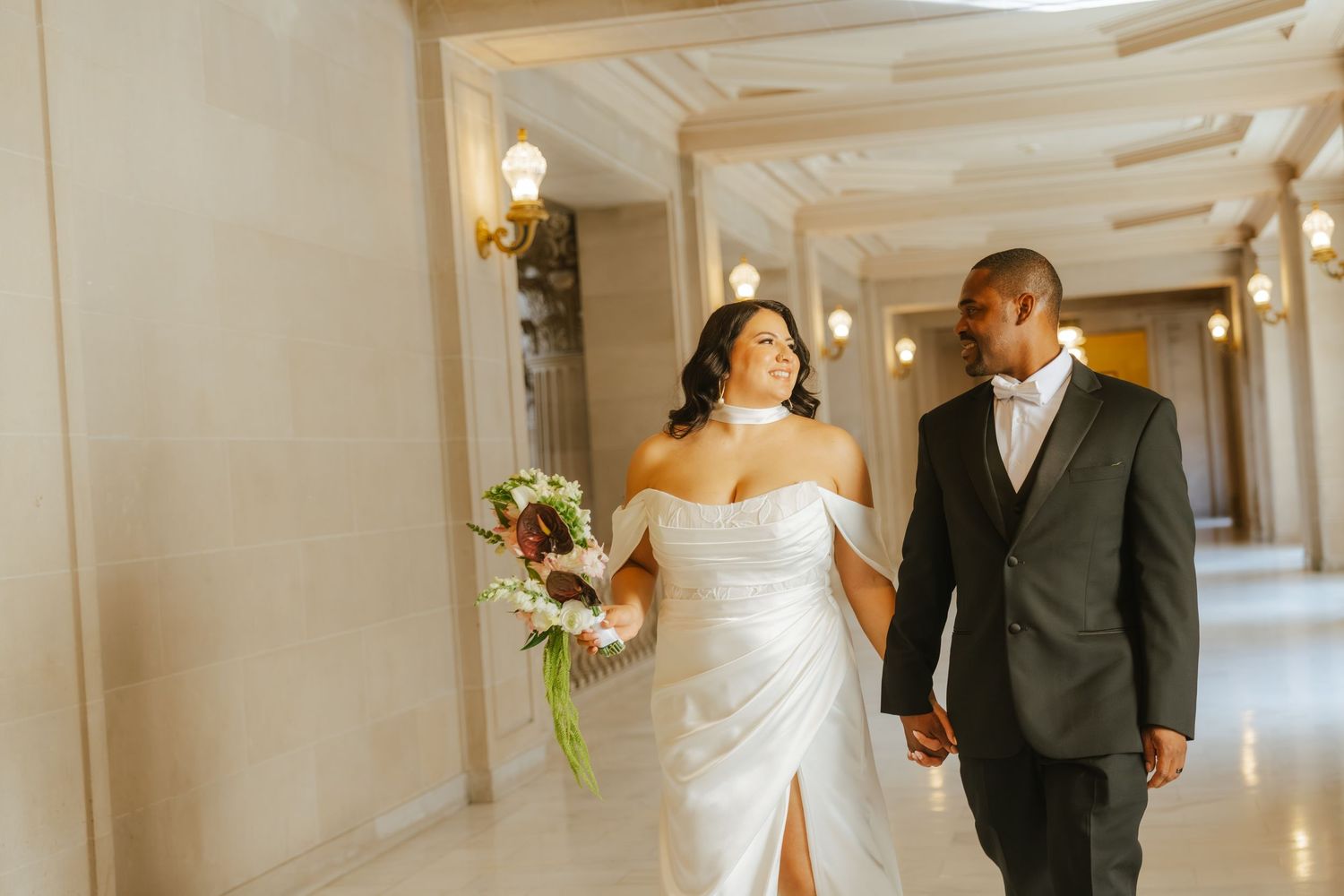 Bride and groom walk hand-in-hand through elegant marble hallway, bride holding bouquet in white off-shoulder gown.