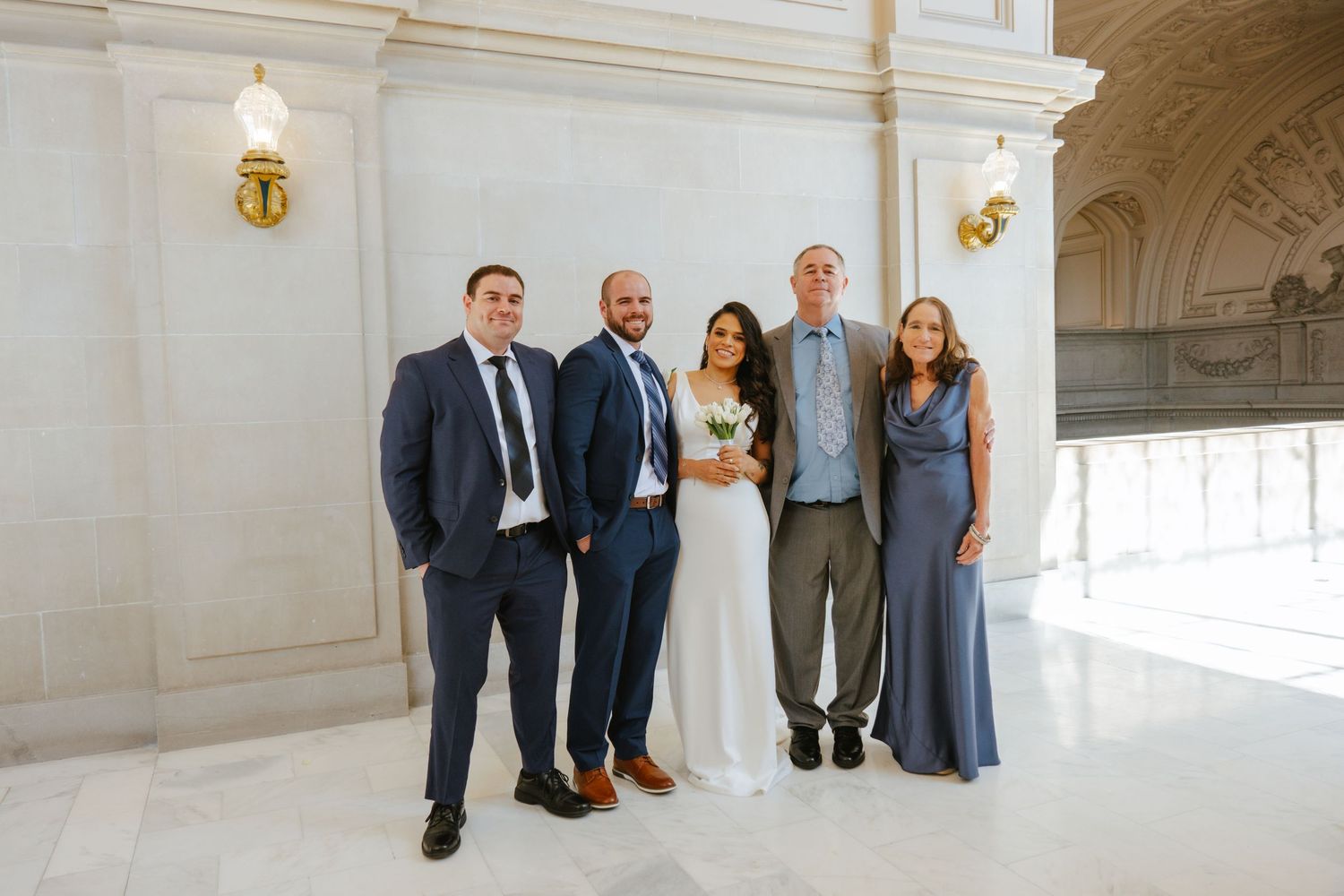 Wedding party members stand together in an elegant white marble hallway with ornate architectural details.