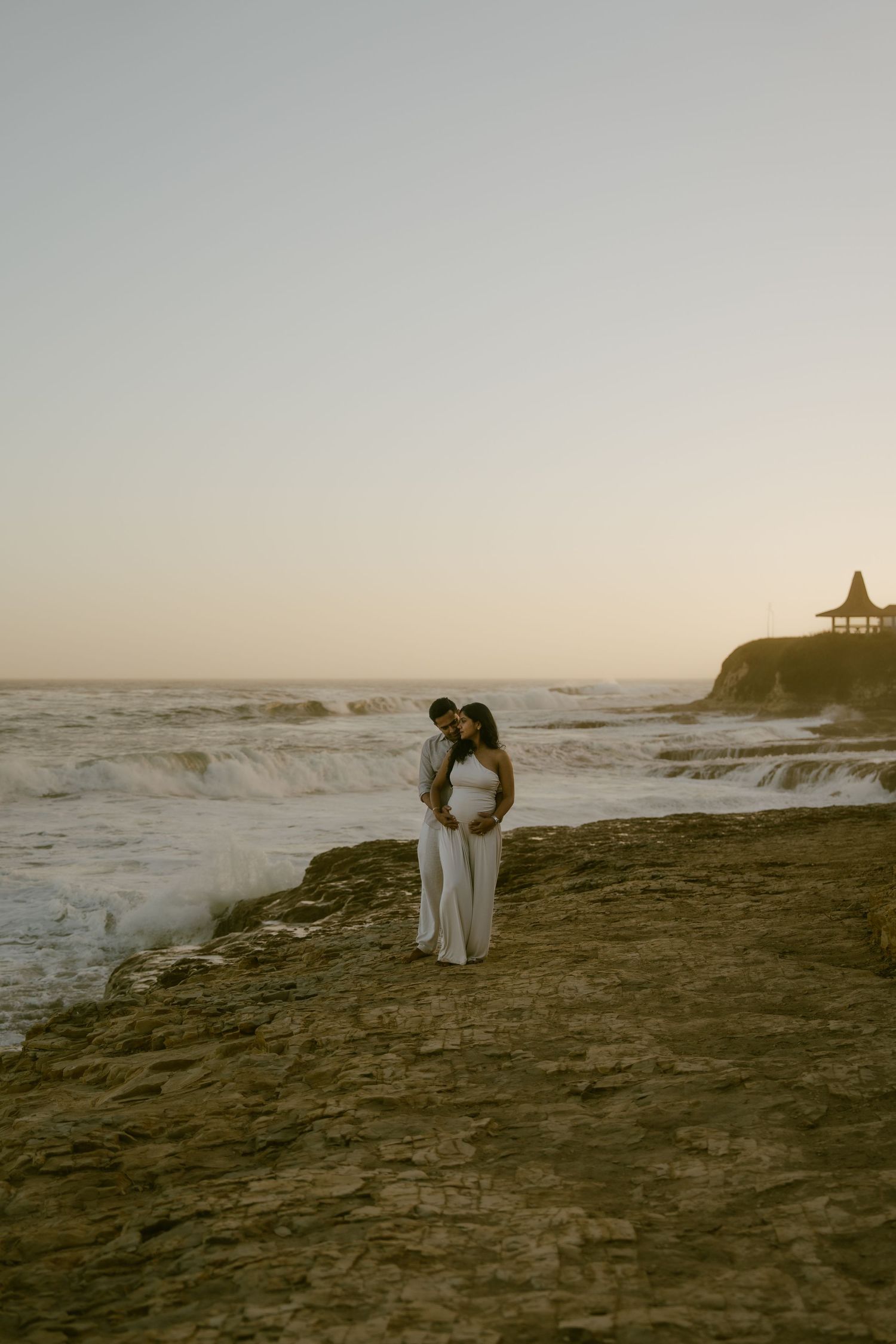 Couple embracing on rocky beach at sunset, woman in white dress with baby bump, ocean waves and cliff gazebo in background.