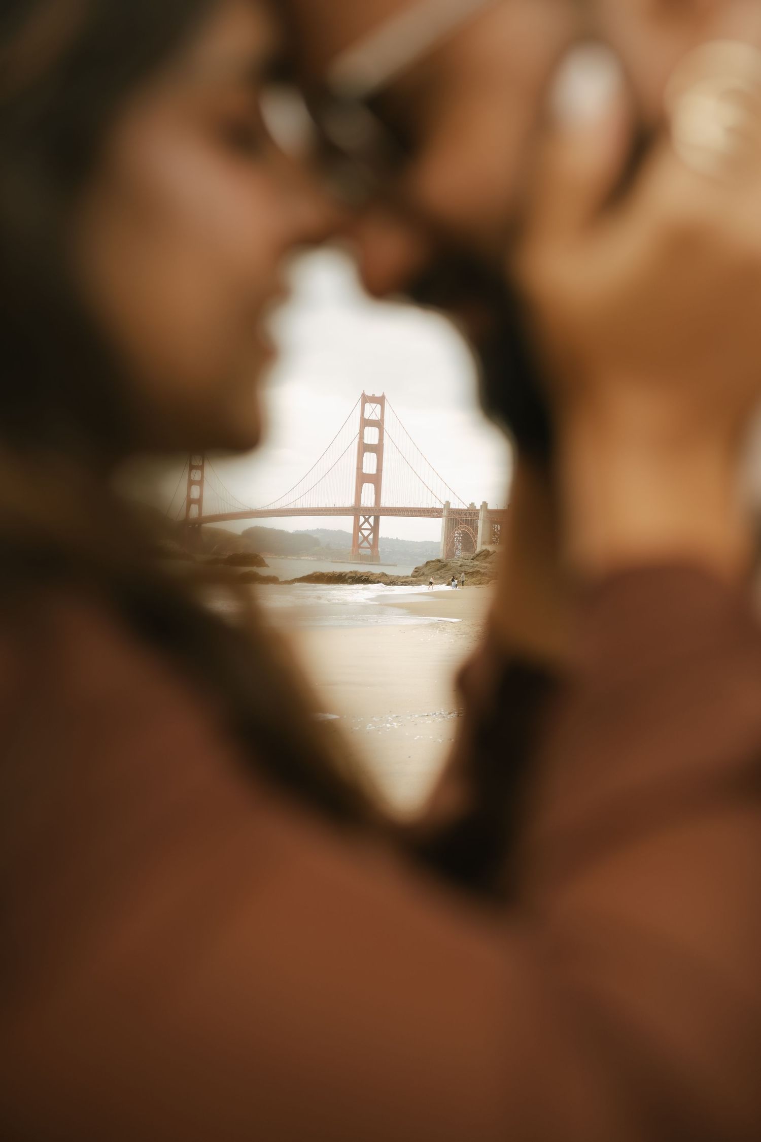 Couple kissing with Golden Gate Bridge framed between them in soft bokeh, San Francisco romantic engagement photo.