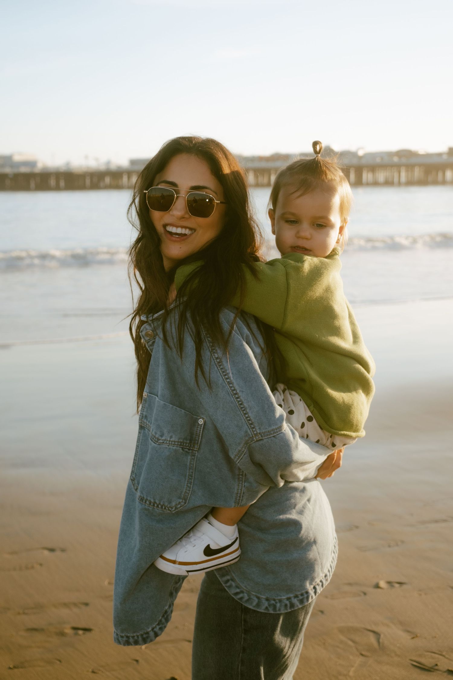 Woman in denim outfit holding toddler on beach, smiling, wearing sunglasses, with ocean and pier in background.