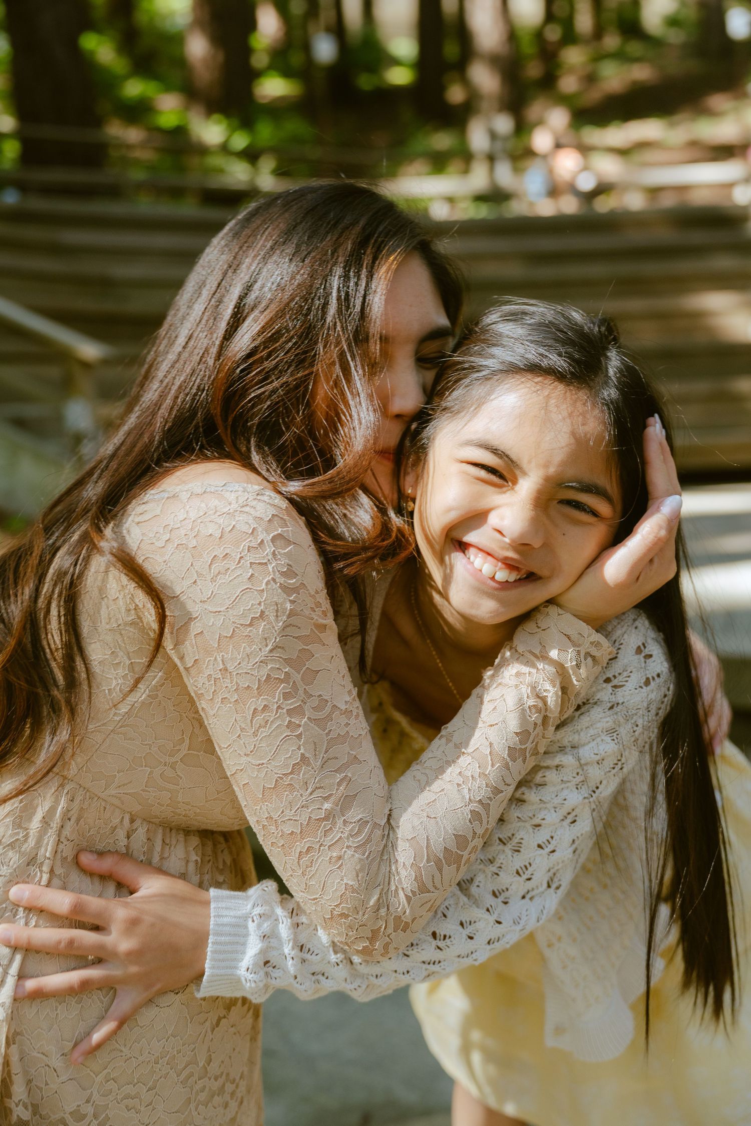 Two young girls hugging and smiling outdoors in cream lace outfits, sharing a joyful moment in a sunny park.