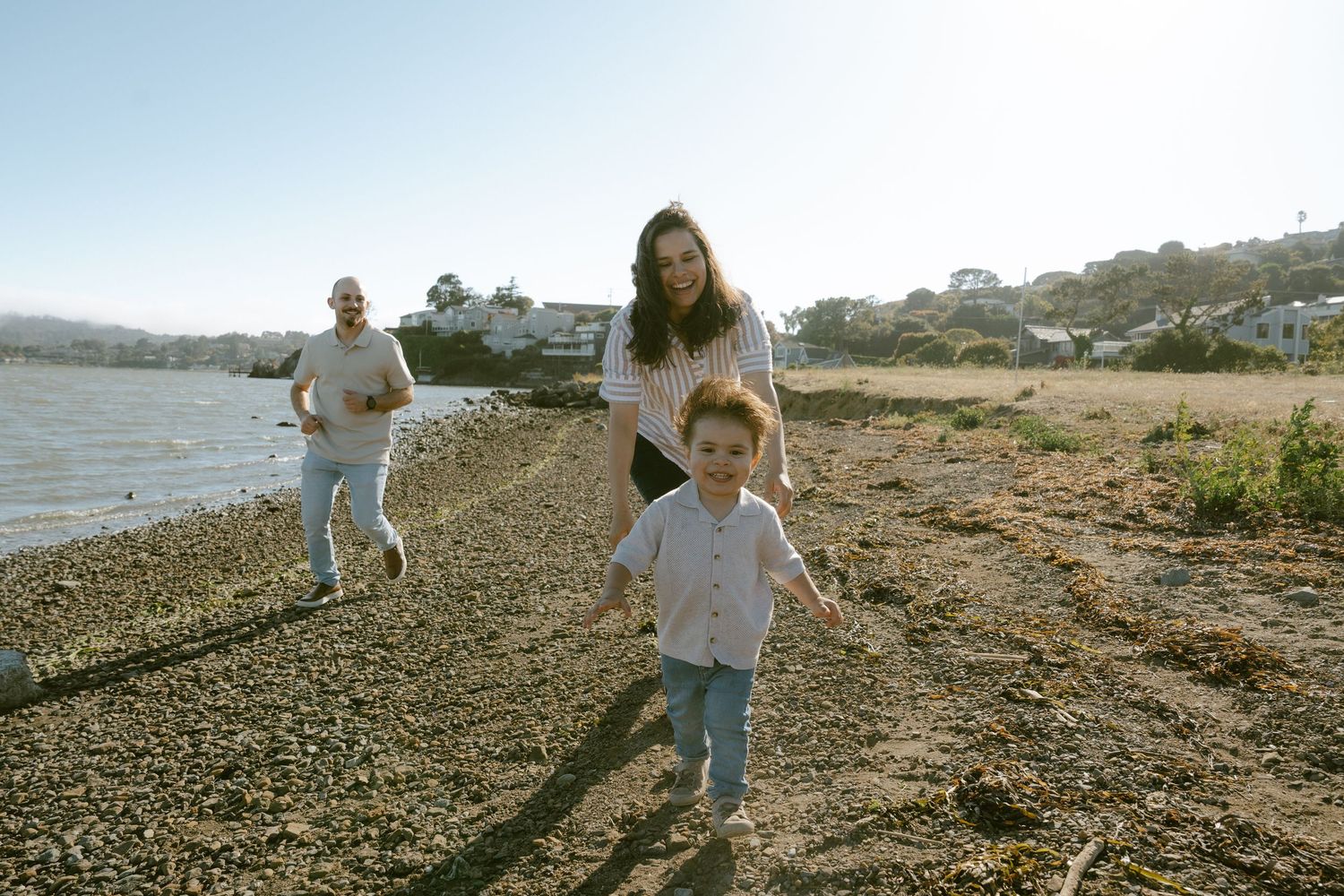 Family enjoying a sunny day on a rocky beach, with a woman and young child running while a man walks behind.