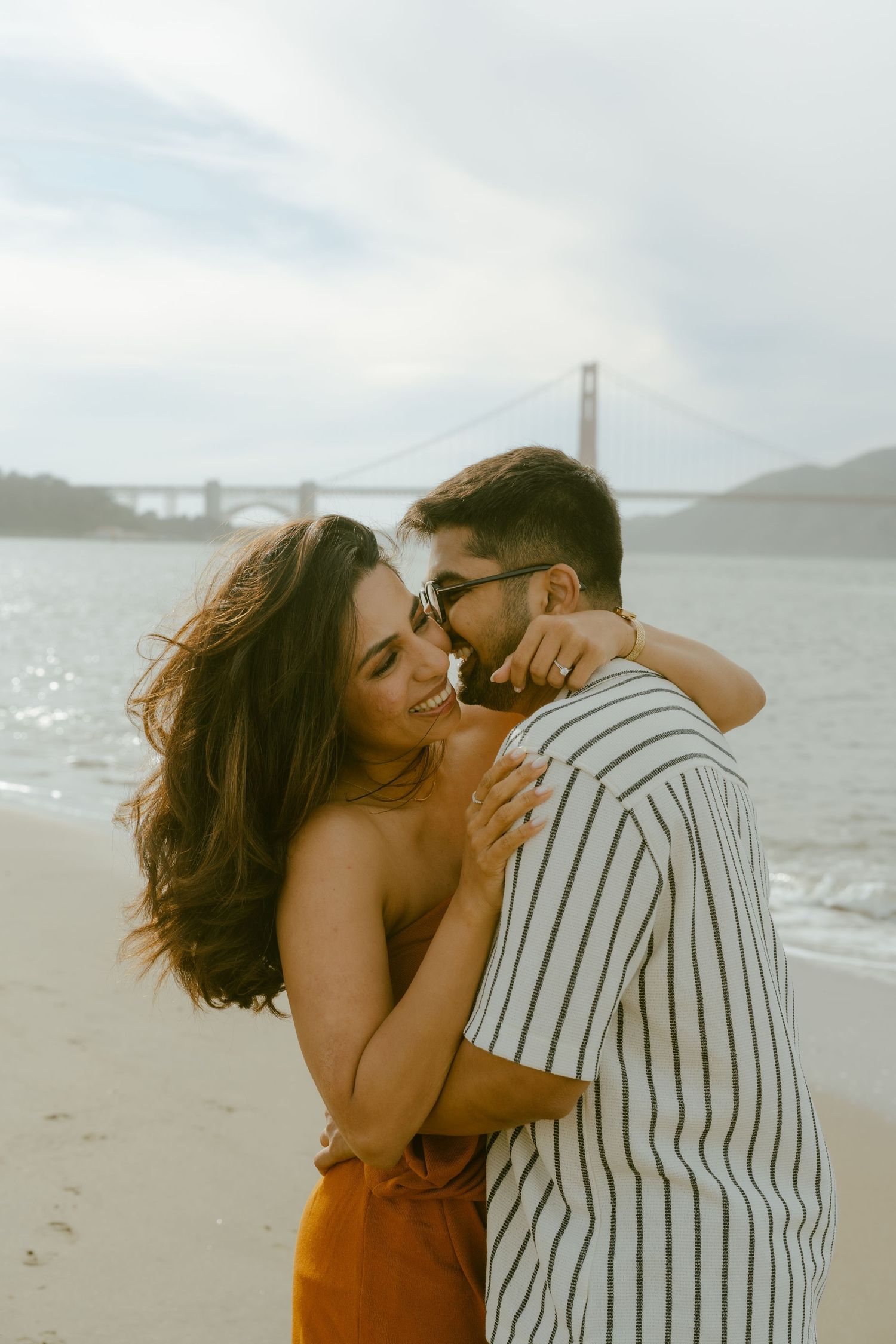 Couple embracing romantically on a beach with the Golden Gate Bridge in the background on a cloudy day.