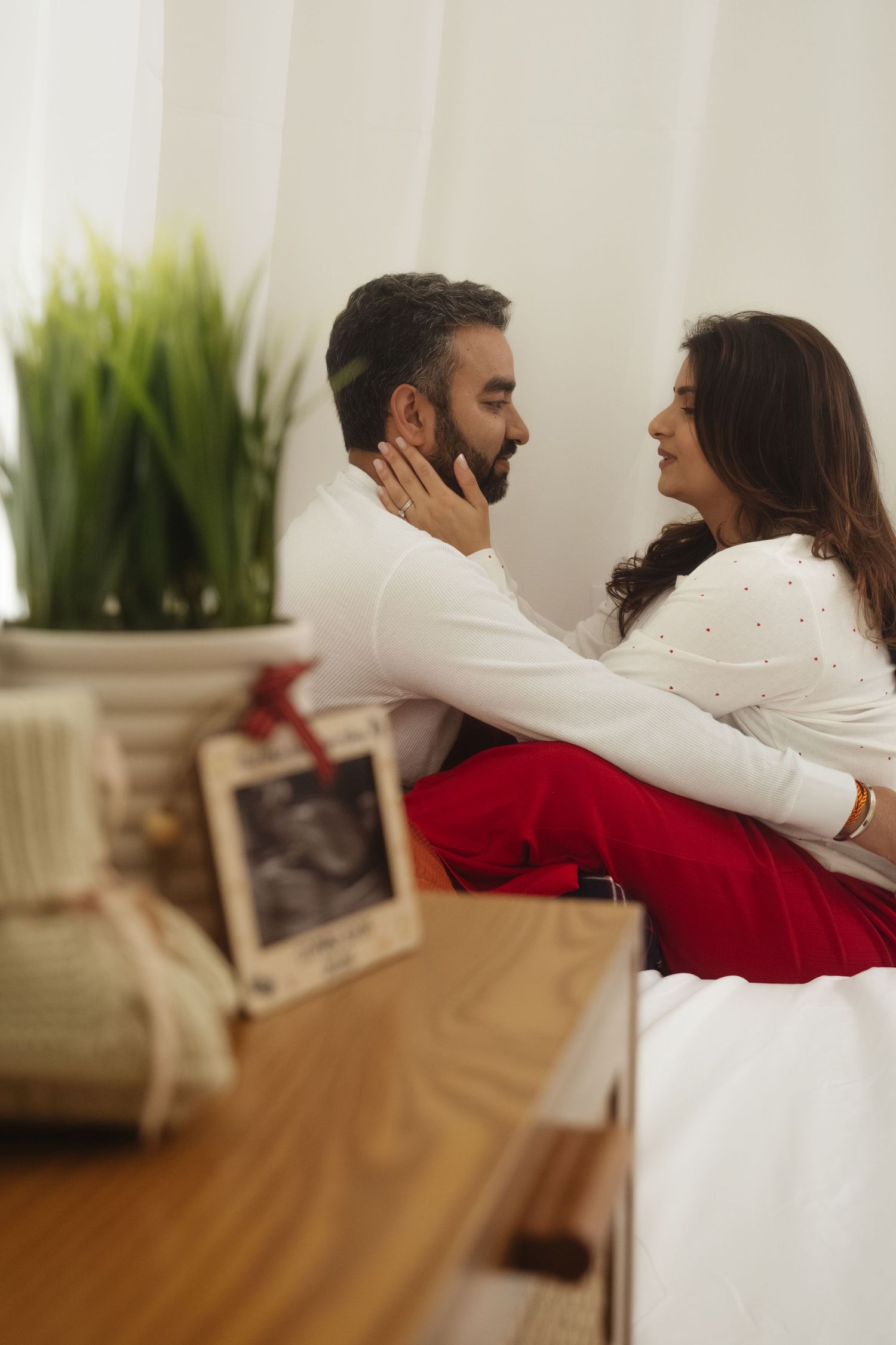 Intimate couple embracing on couch in bright living room with white curtains and plants.